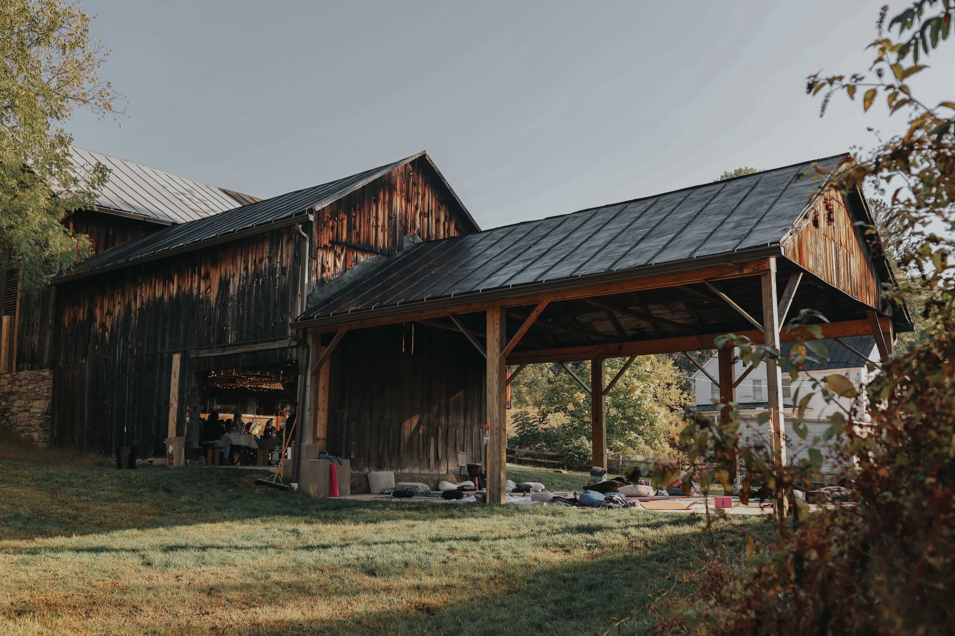 A rustic wooden barn with a metal roof stands next to an open shelter on a grassy area.