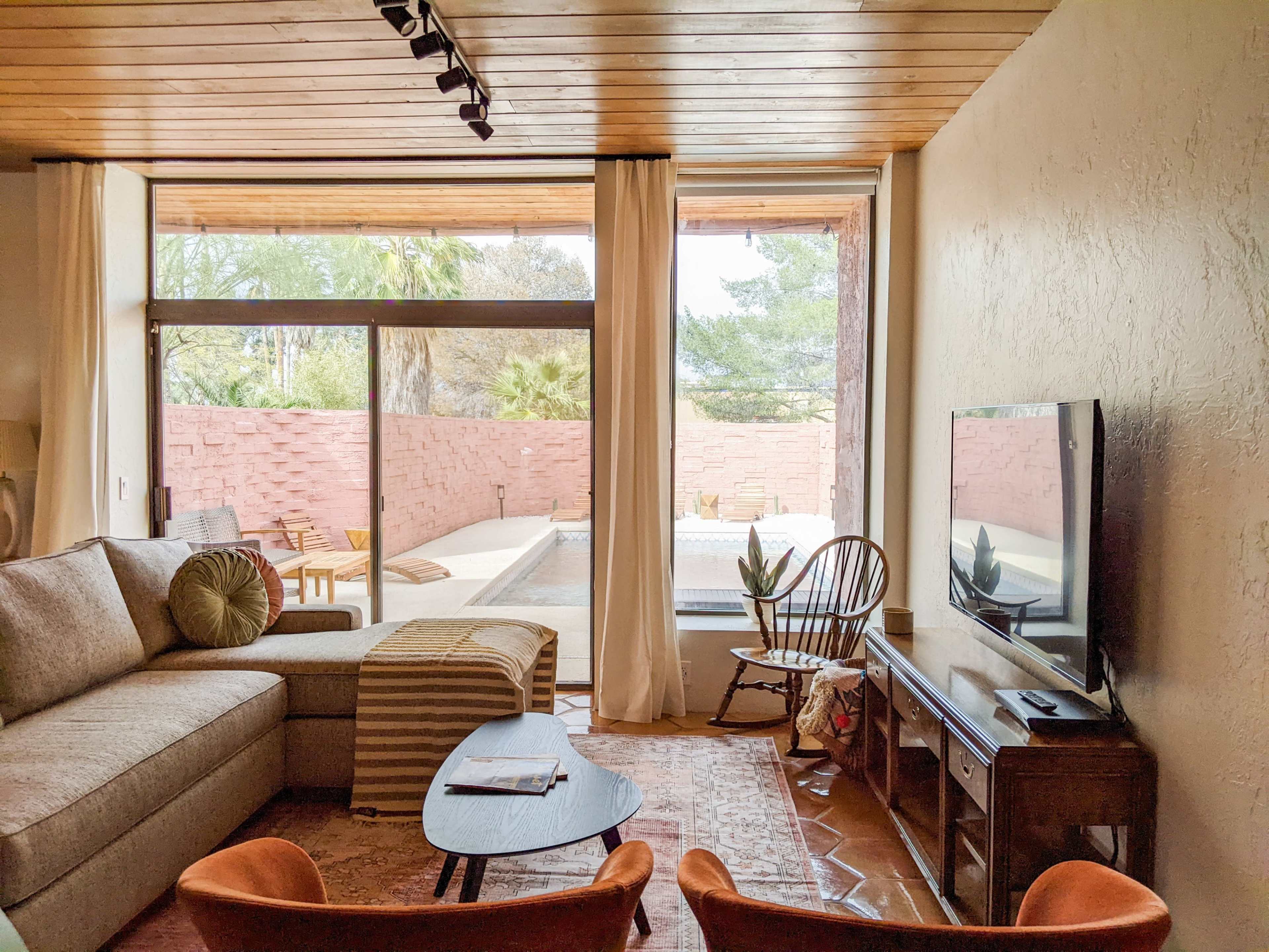 A modern living room with a large window overlooking a patio and pool area, featuring a sectional sofa, a coffee table, and a television.