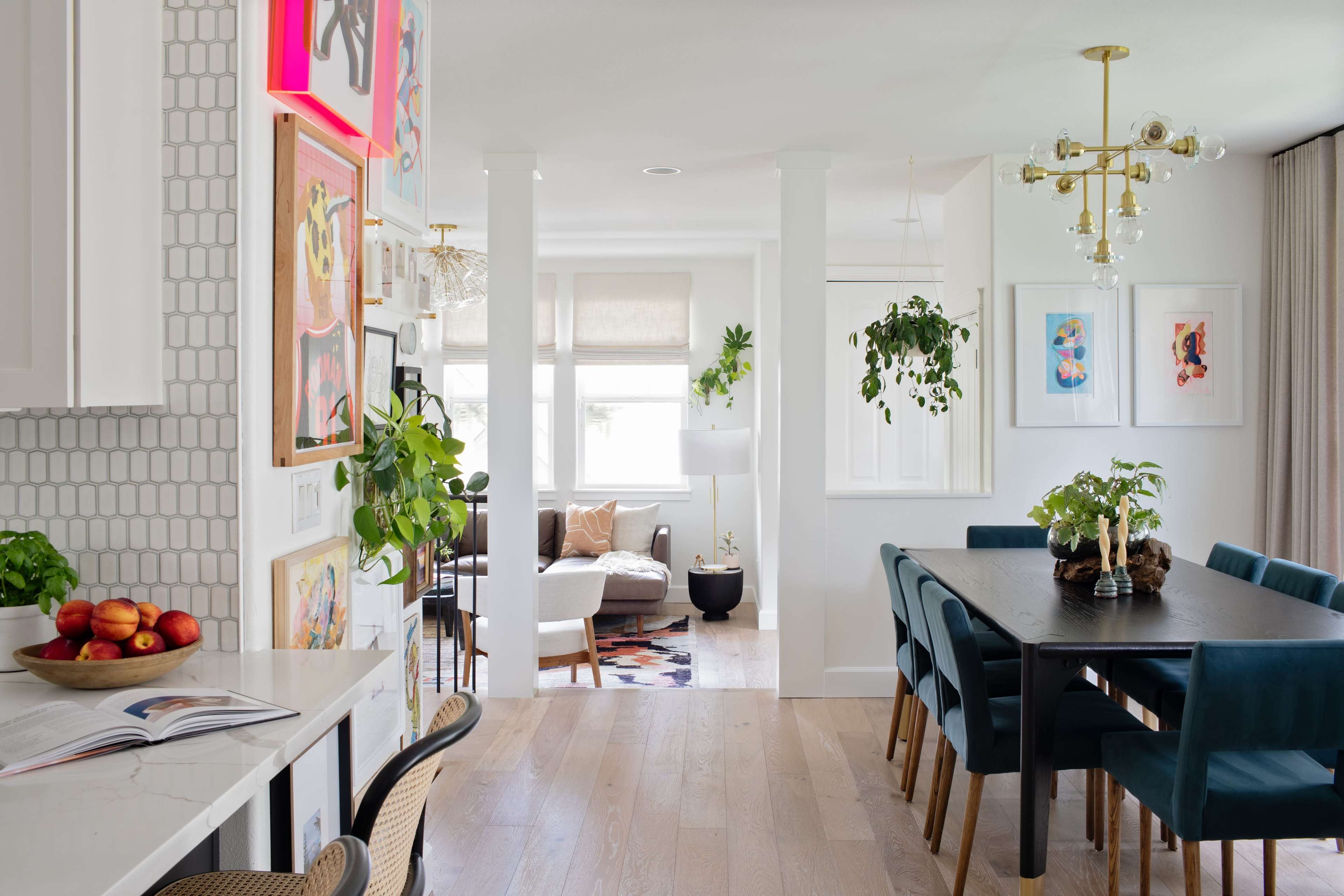 A modern open-concept living space featuring a dining area with a large table, stylish chairs, a gallery wall of framed artwork, and plants, leading into a cozy living room.