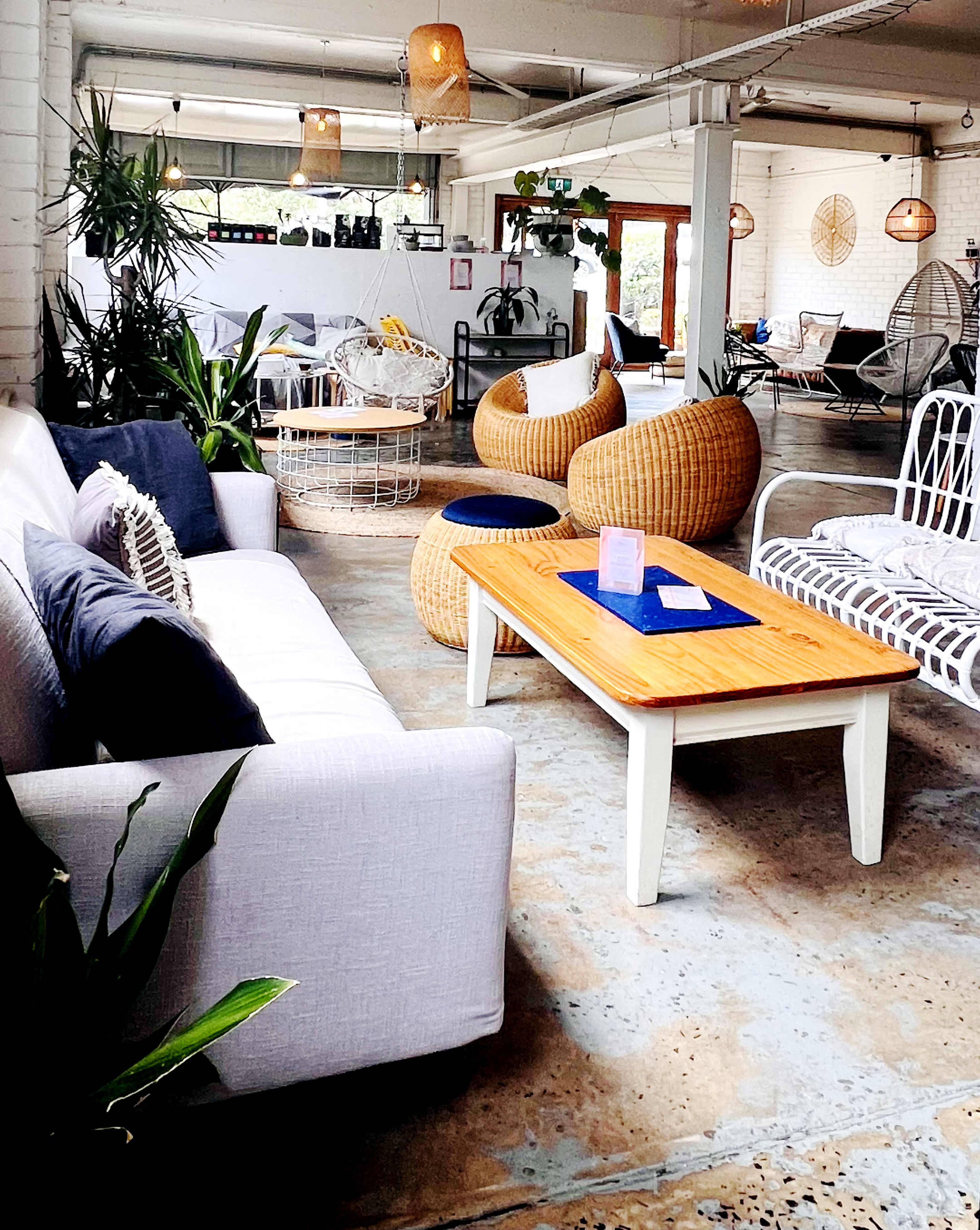 The image shows a modern indoor space featuring a white sofa, woven armchairs, a wooden coffee table, and various plants.