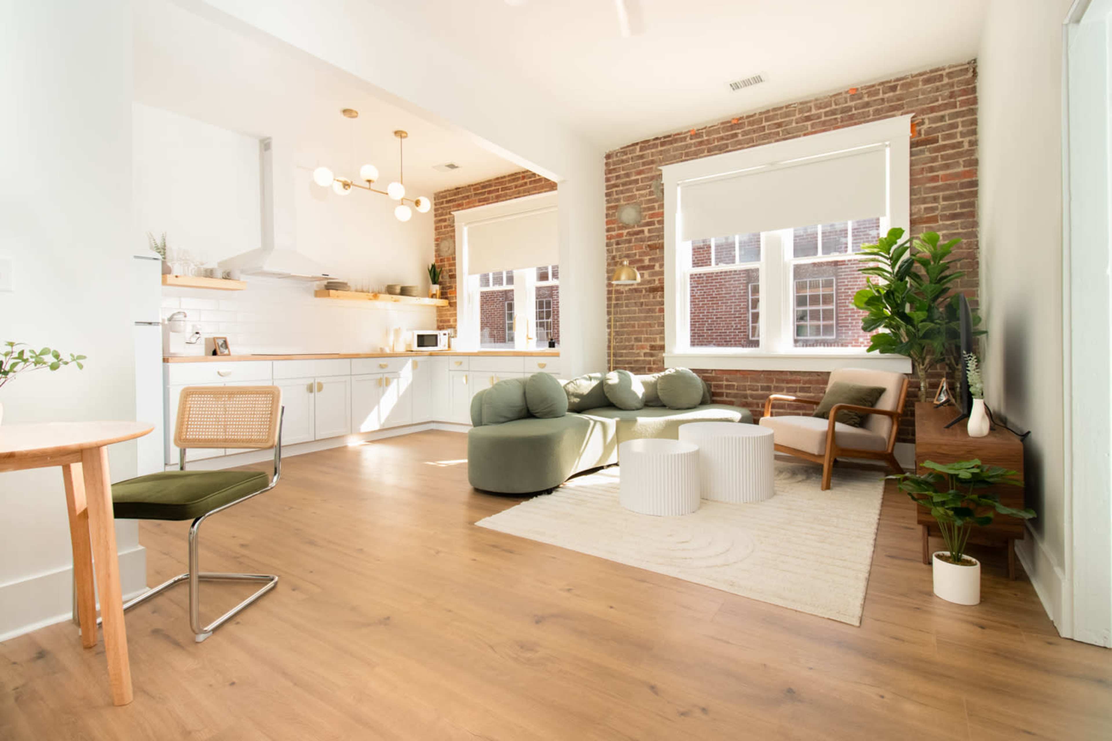 A modern living space with a kitchen area, featuring a seating arrangement of round chairs and a coffee table, alongside large windows and exposed brick walls.