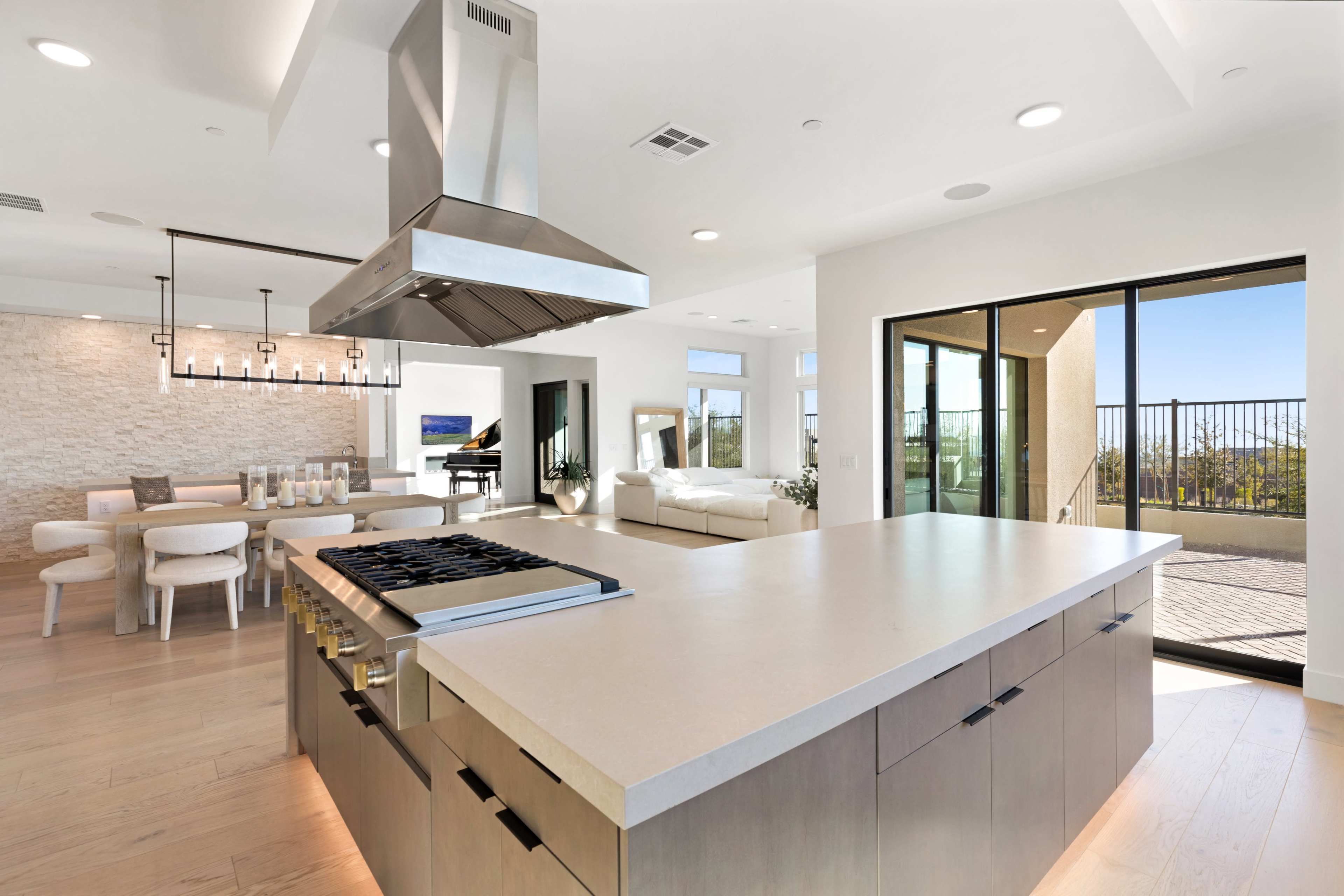 A modern kitchen with a central island, stainless steel range hood, and an open layout that connects to a dining area and a balcony with a view.