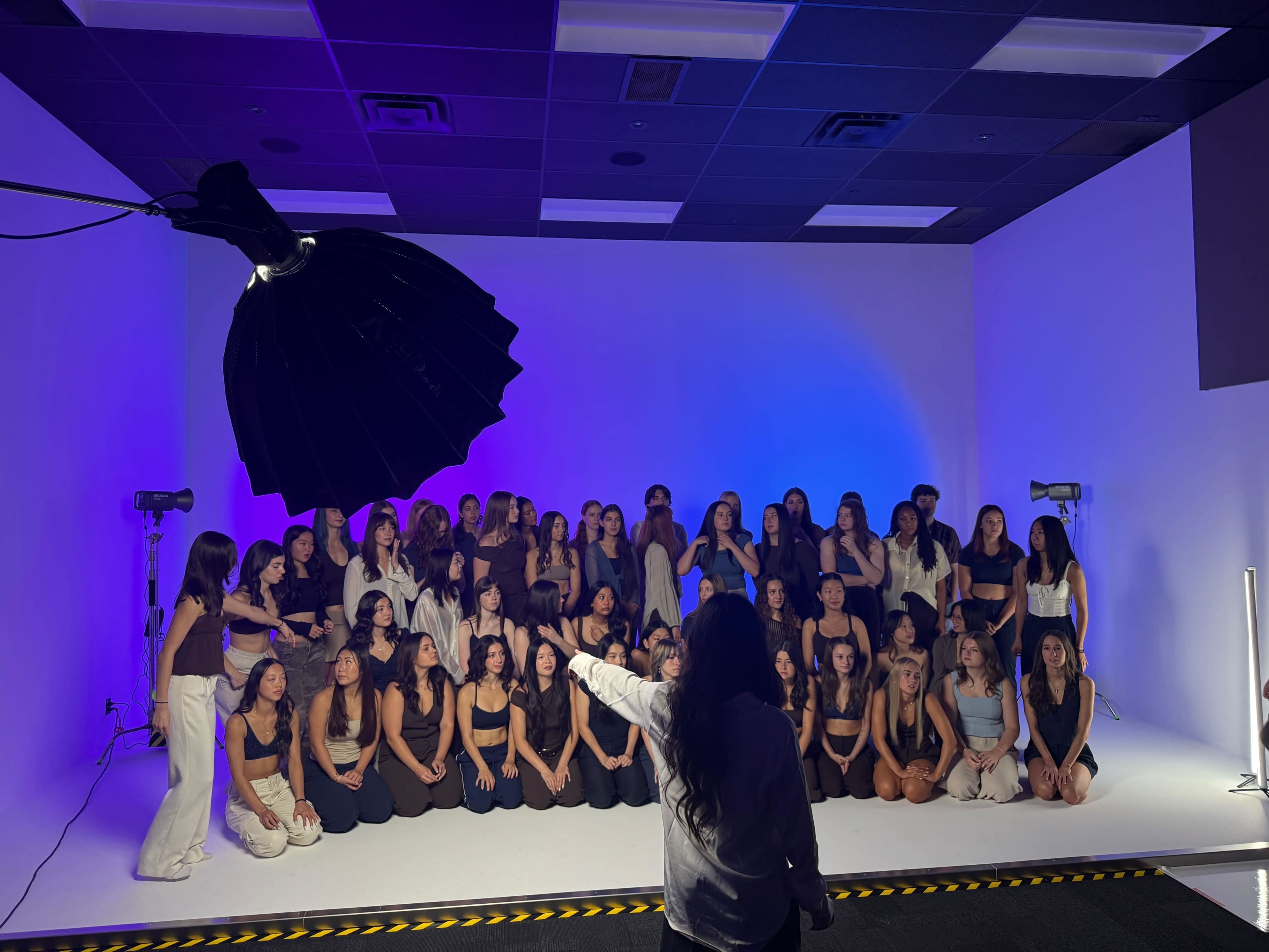 A large group of women is posed for a photoshoot in a studio with colorful lighting and professional equipment.