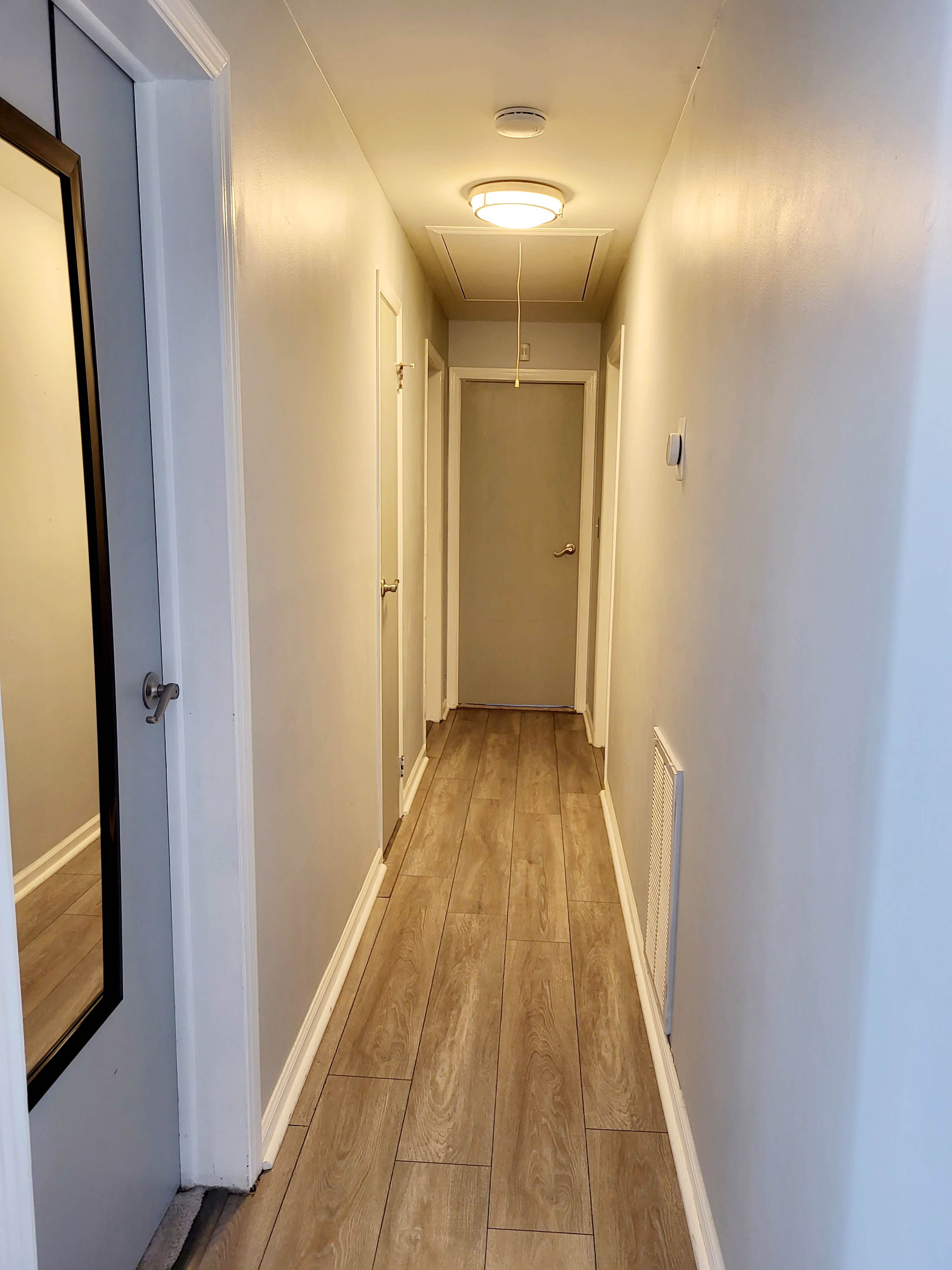 A narrow hallway with light-colored walls, a mirror on the left, and a closed door at the end.