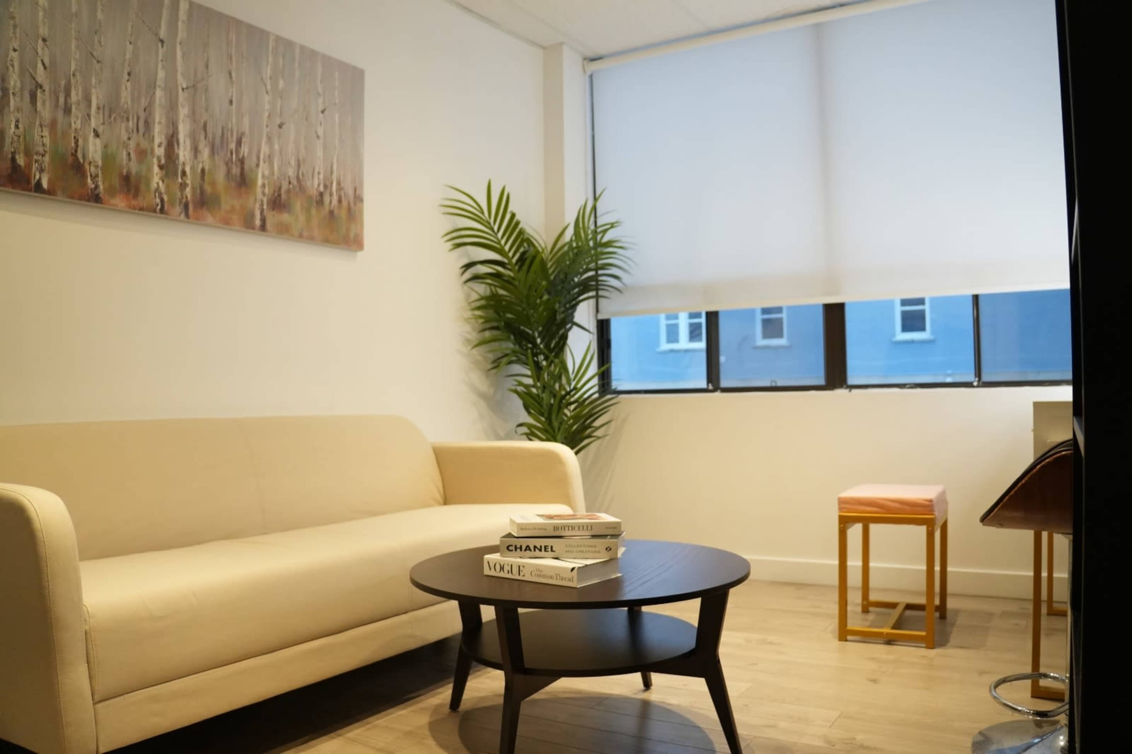 The image shows a modern waiting area with a beige couch, a round black coffee table stacked with magazines, a potted plant, and a window with partially drawn blinds.
