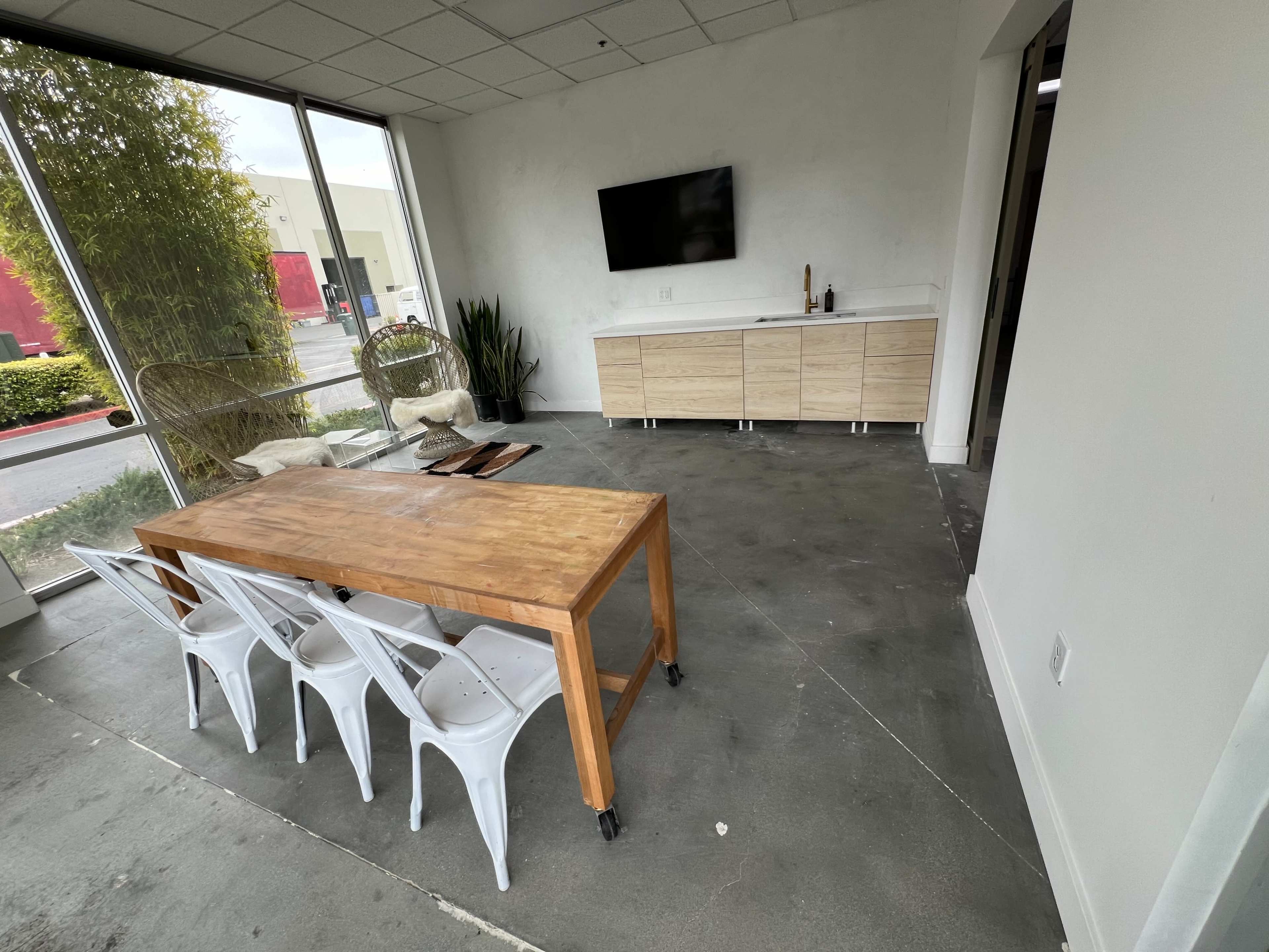 The image shows a modern, minimalistic interior with a wooden table and four white chairs, a sideboard, a television mounted on the wall, and large windows letting in natural light.