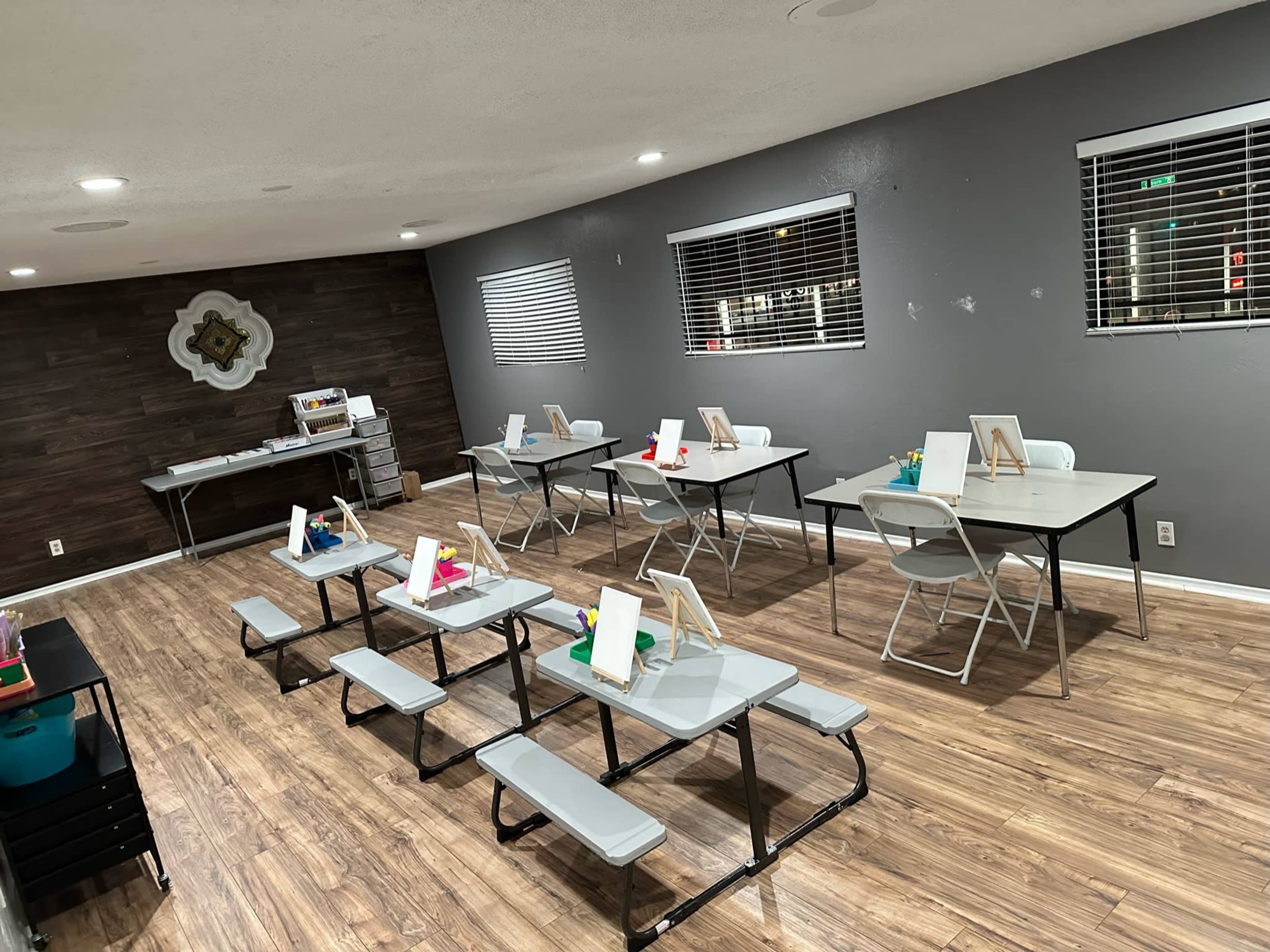 A neatly arranged room with several tables and chairs set up for painting activities, featuring easels and supplies at each place.