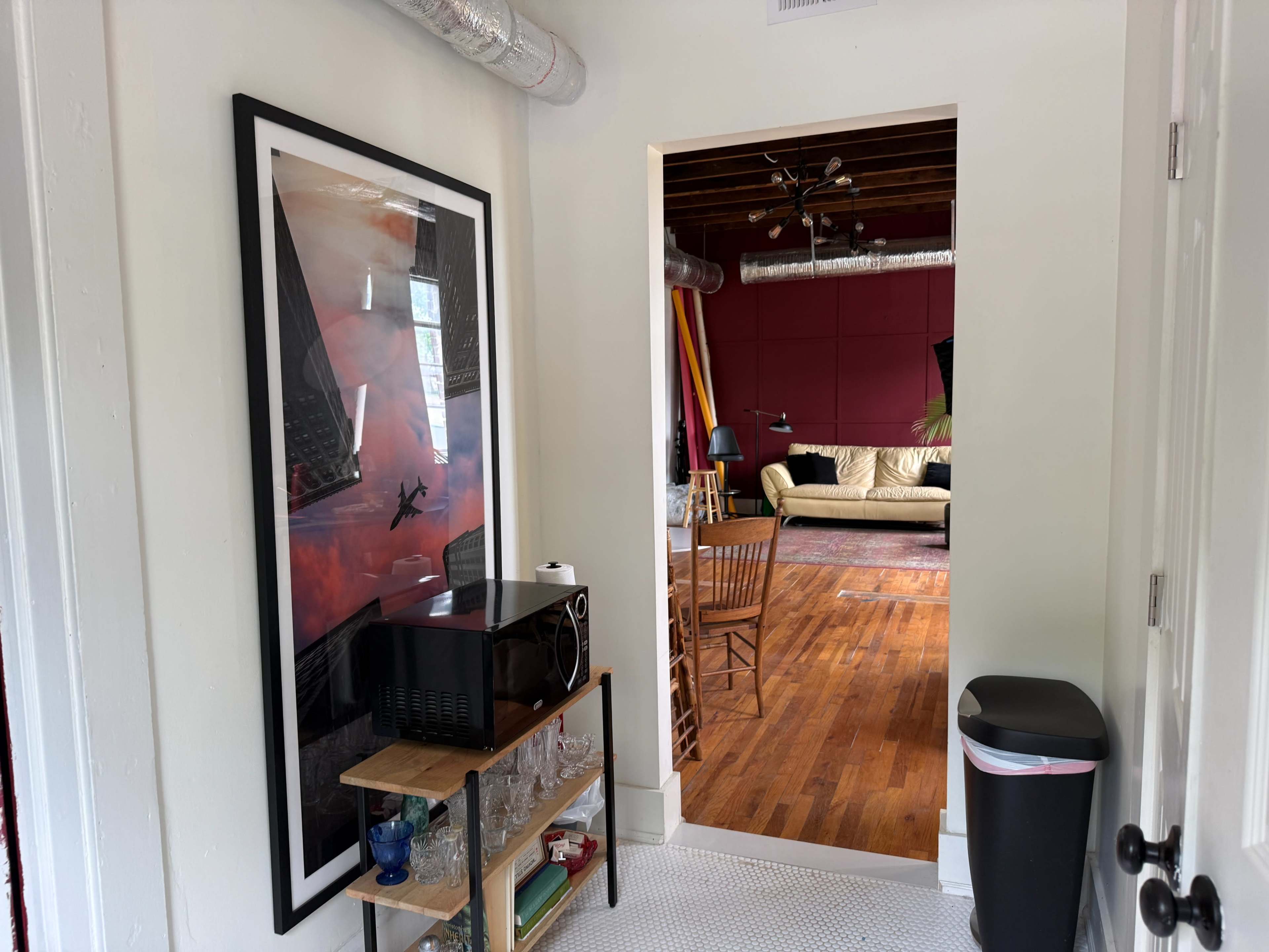 The image shows a hallway with a large framed poster, a microwave on a wooden cart, and a view into a room featuring a sofa, colorful walls, and wooden flooring.