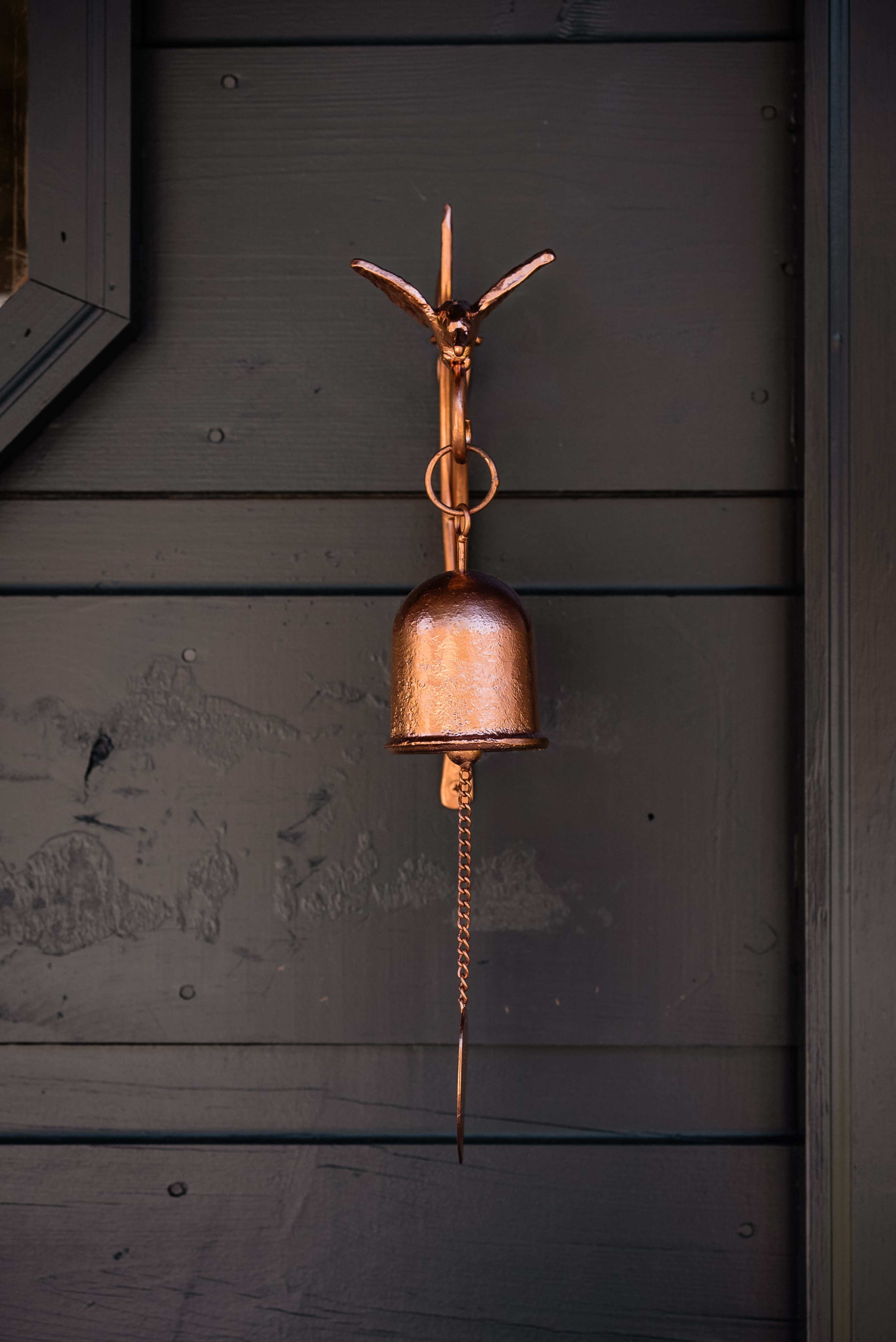 A copper bell with a decorative mount and a chain hangs on a dark wooden wall.