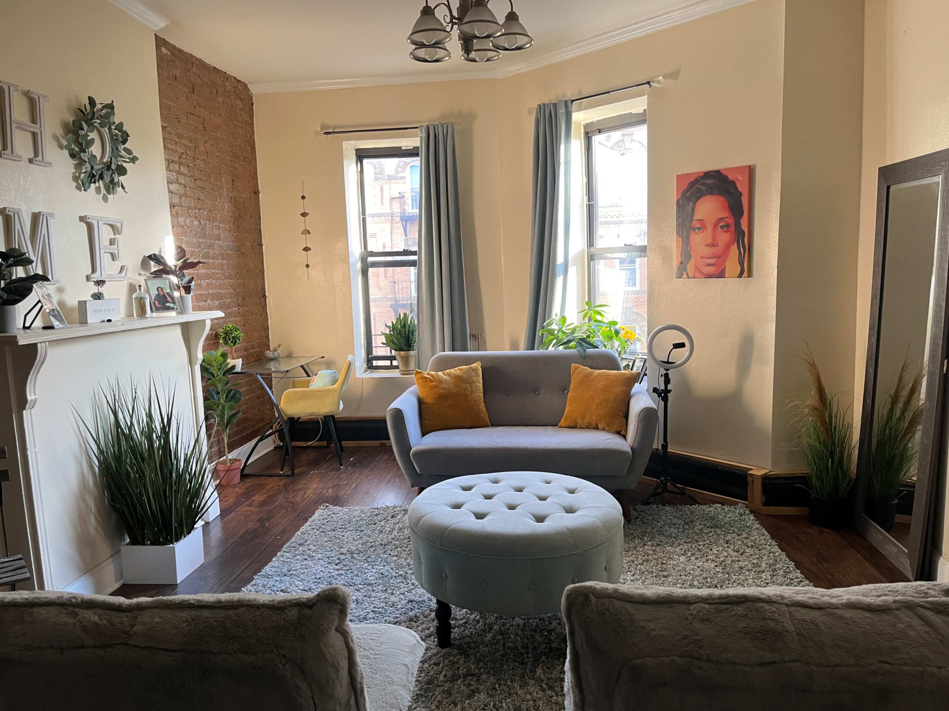 A cozy living room with a light gray sofa, a round ottoman, and a large mirror, featuring decorative plants and a portrait on the wall.
