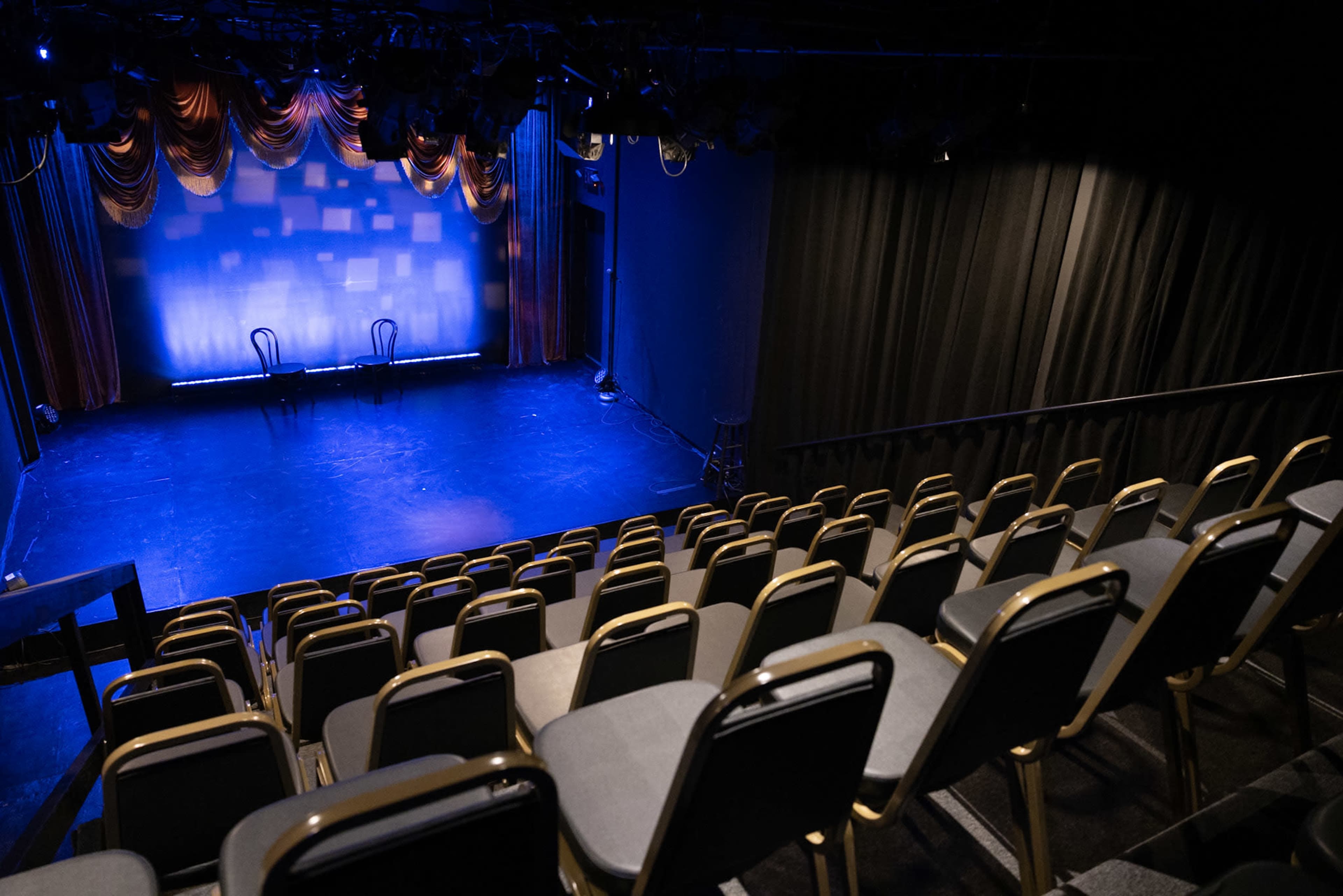 A small theater stage with two chairs on it, surrounded by empty rows of seating and dramatic curtains.