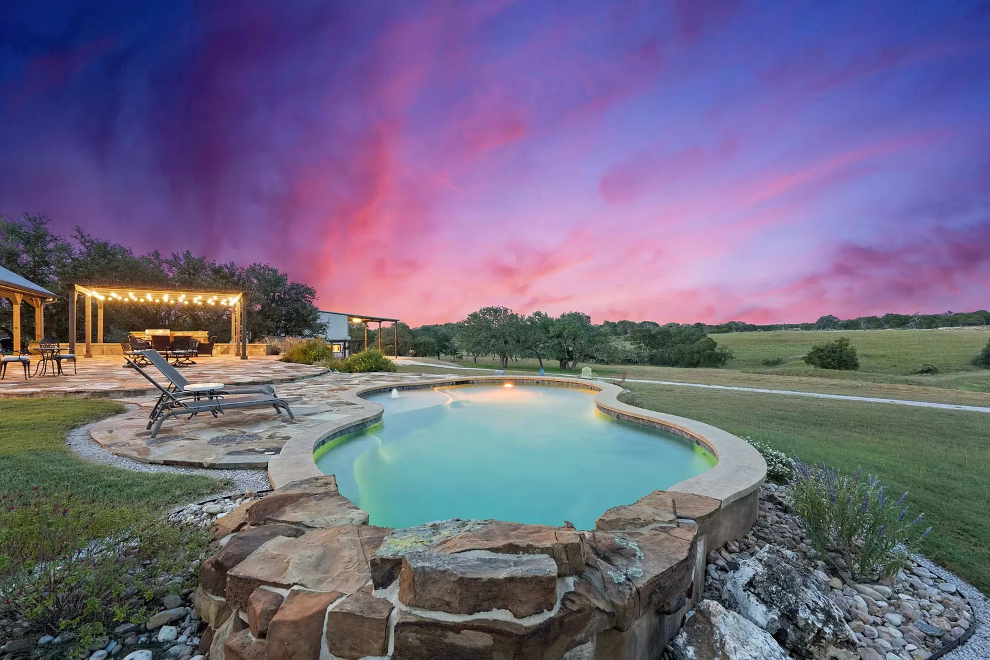 A serene pool with stone edging reflects a colorful sunset sky, surrounded by lush green fields and trees.