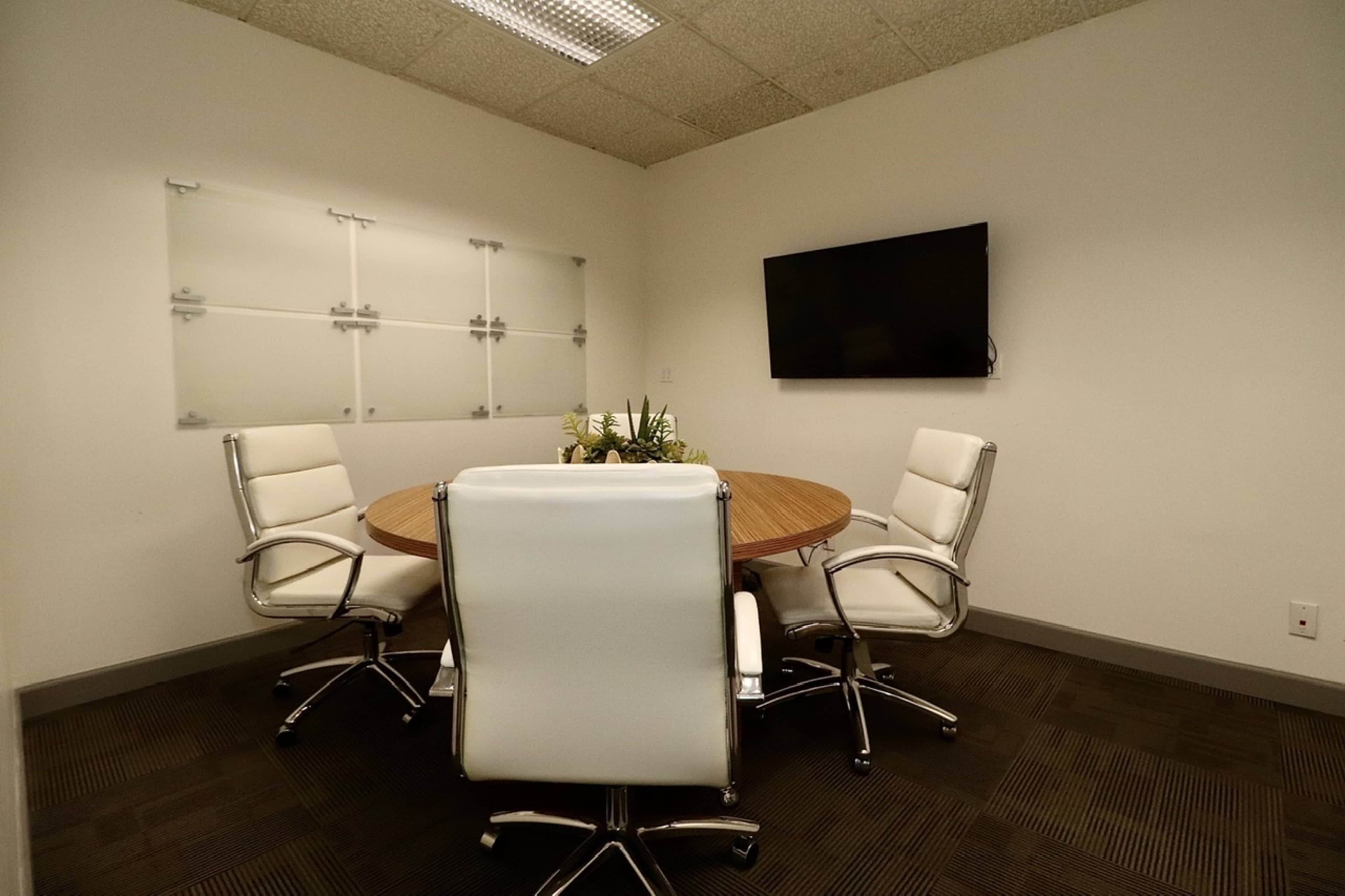 A small conference room features a round wooden table surrounded by four white leather chairs, with a mounted television and a glass whiteboard on the wall.
