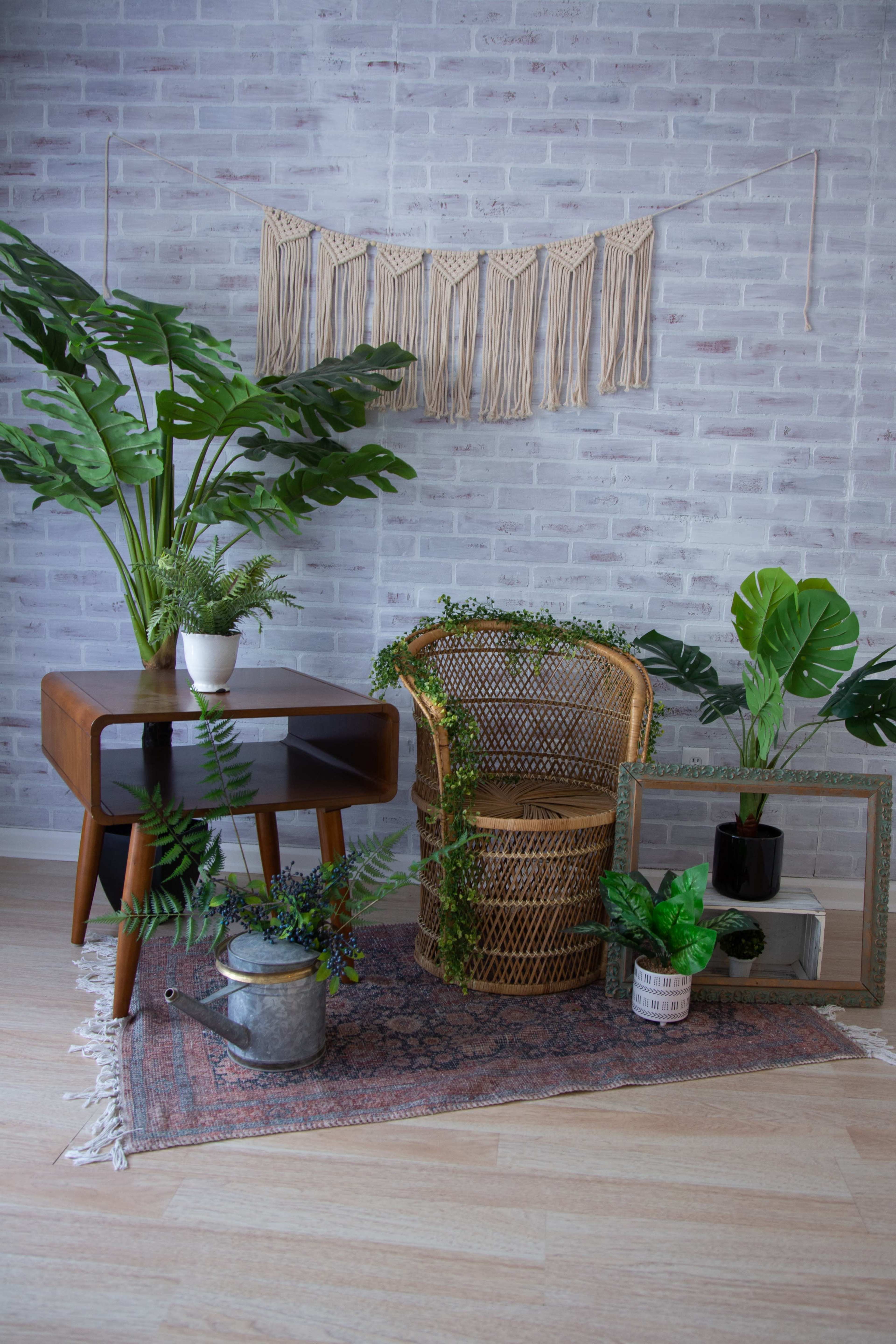 A cozy interior with a rattan chair, a wooden table, and various potted plants arranged on a patterned rug against a brick wall.