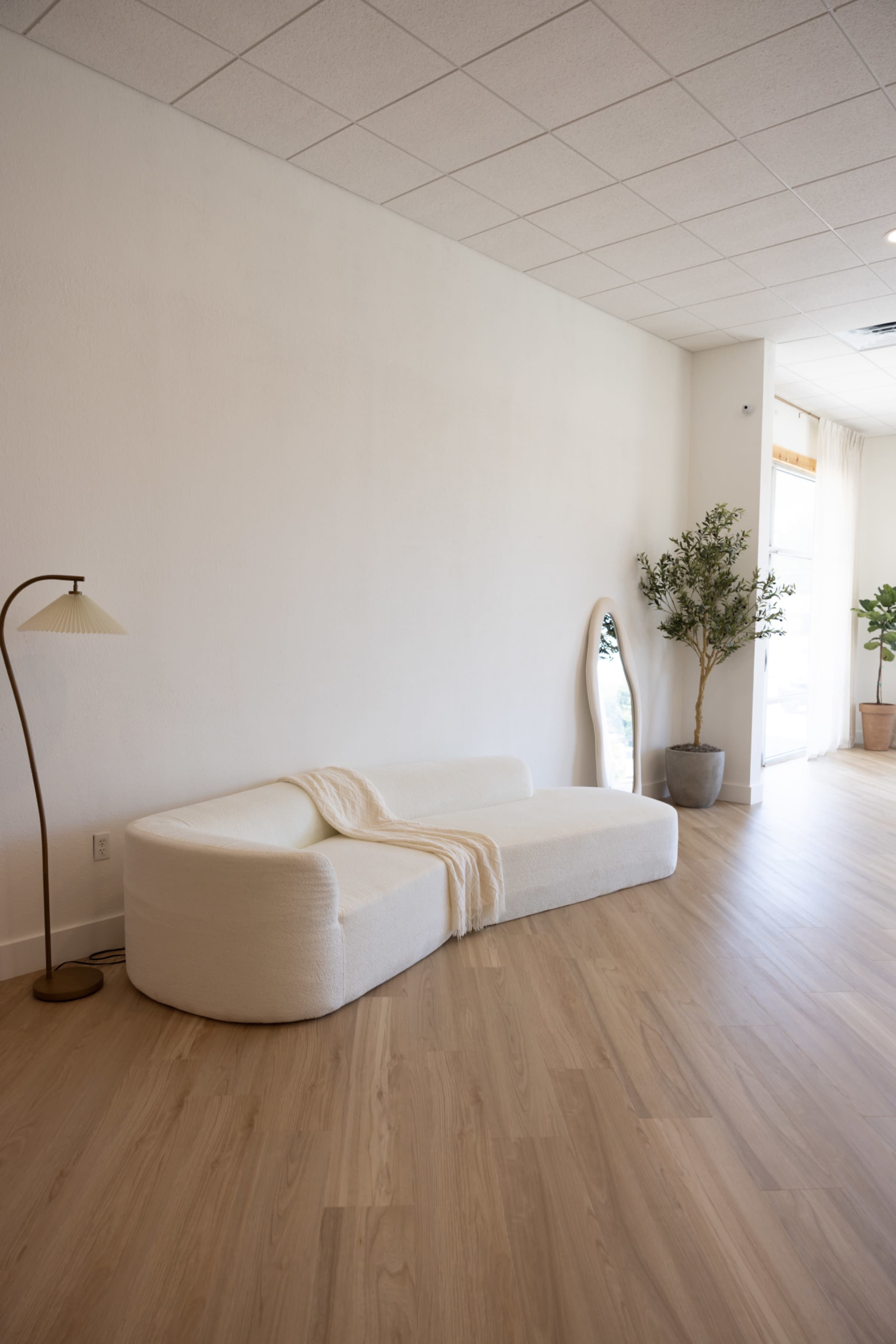 The image shows a minimalist interior space featuring a white curved sofa, a floor lamp, a large mirror, and potted plants on a light wood floor.