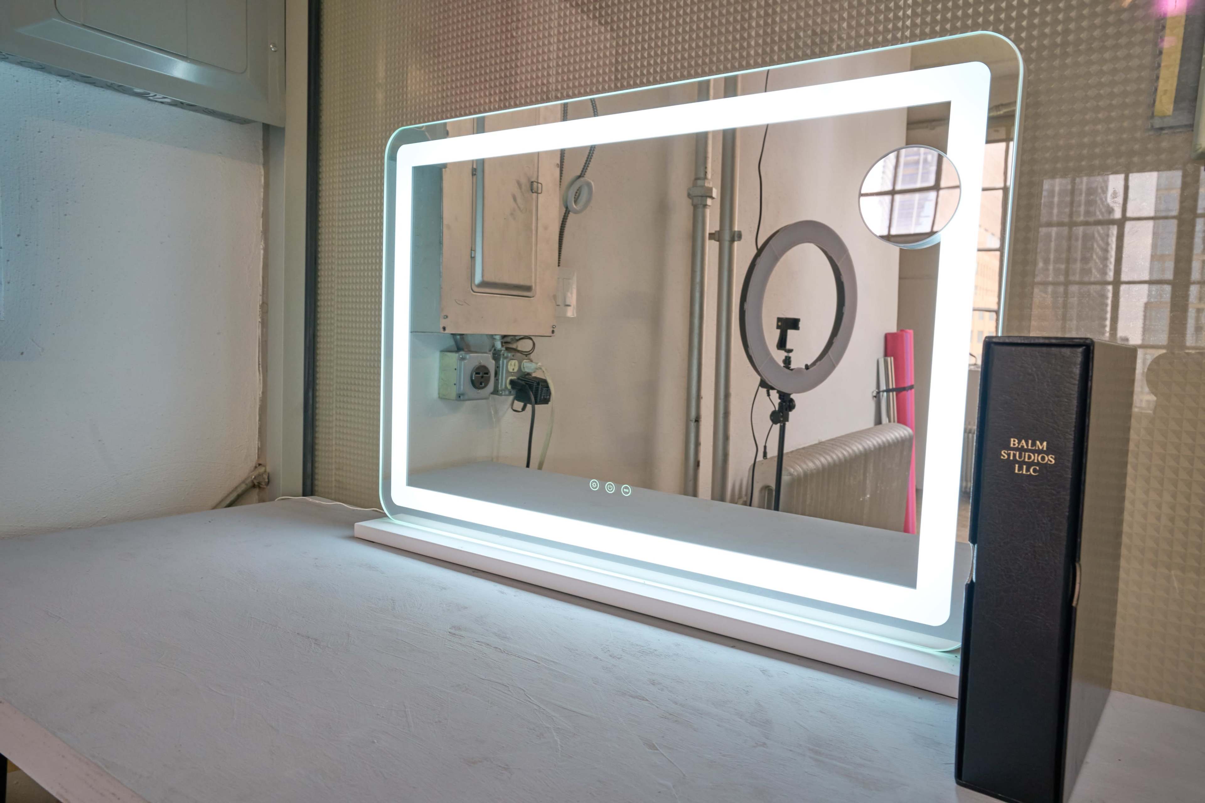 A backlit mirror stands on a white surface, next to a black folder labeled "BALM STUDIOS LLC."