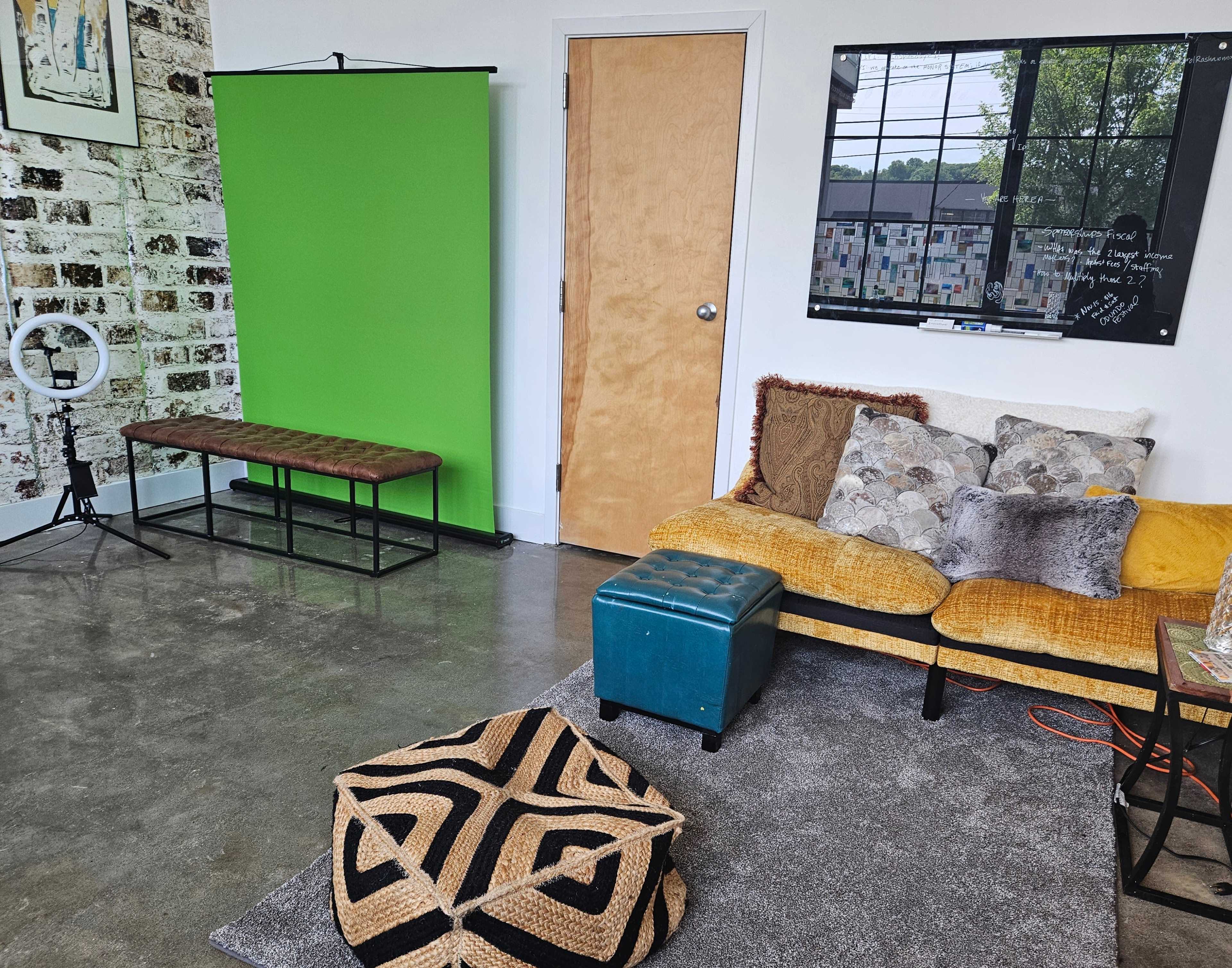 A room with a green backdrop, a bench, a sofa with cushions, a blue ottoman, and a patterned pouf on a rug, along with a window displaying a view outside.