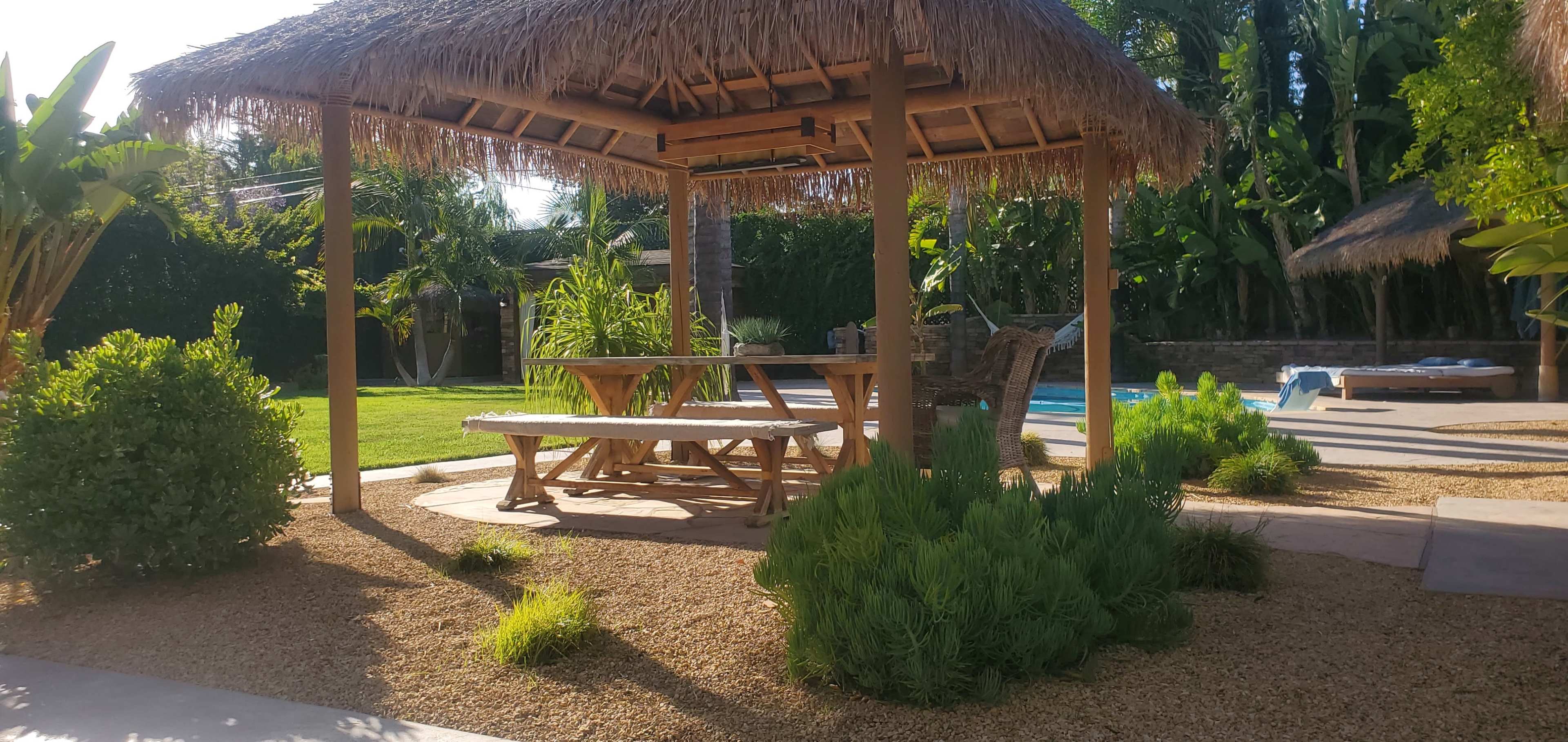 A wooden gazebo with a thatched roof and a picnic table is nestled among landscaped greenery and a swimming pool in the background.