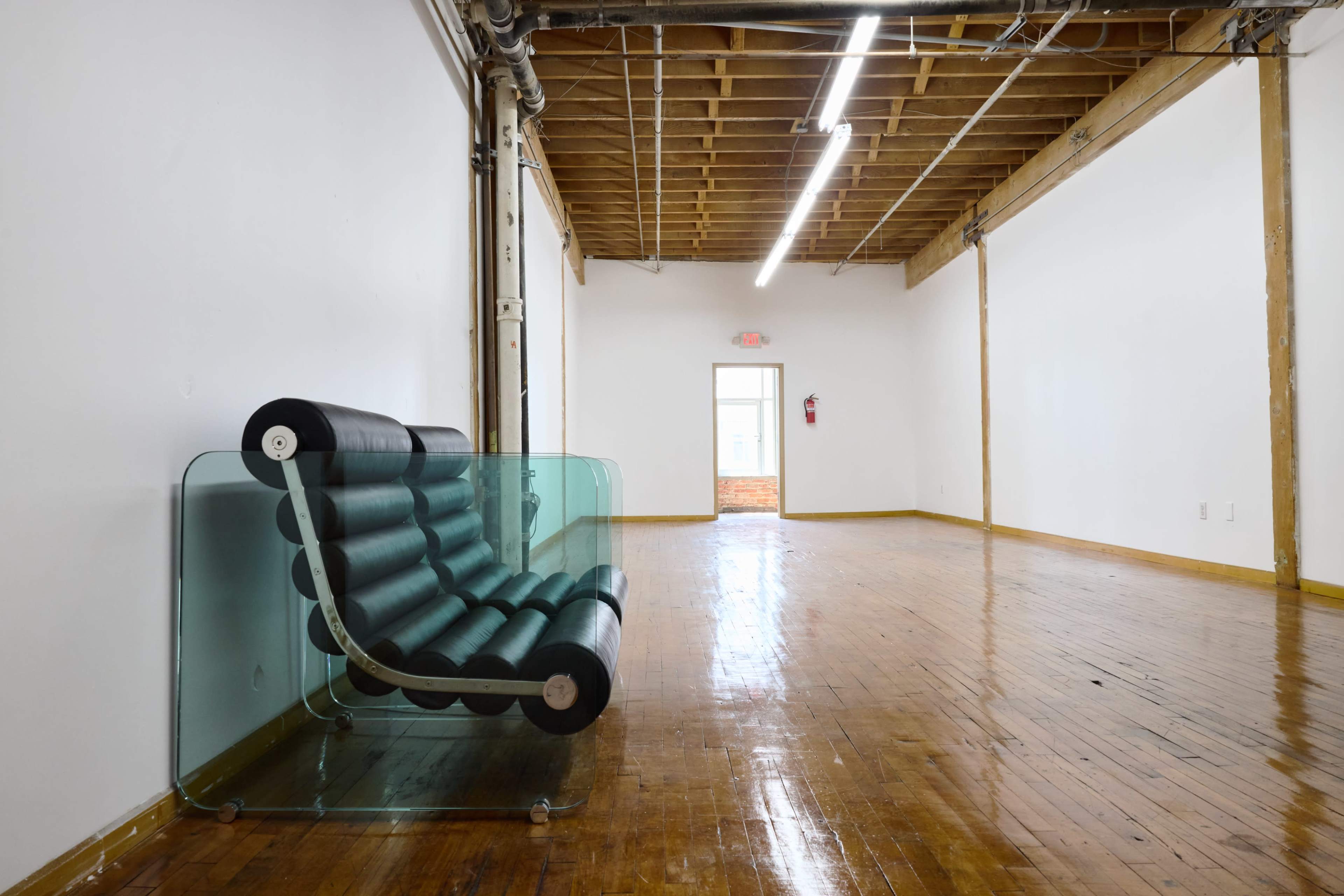 The image shows a spacious, empty room with hardwood floors, featuring a modern black and clear acrylic sofa along one wall and a doorway at the end.