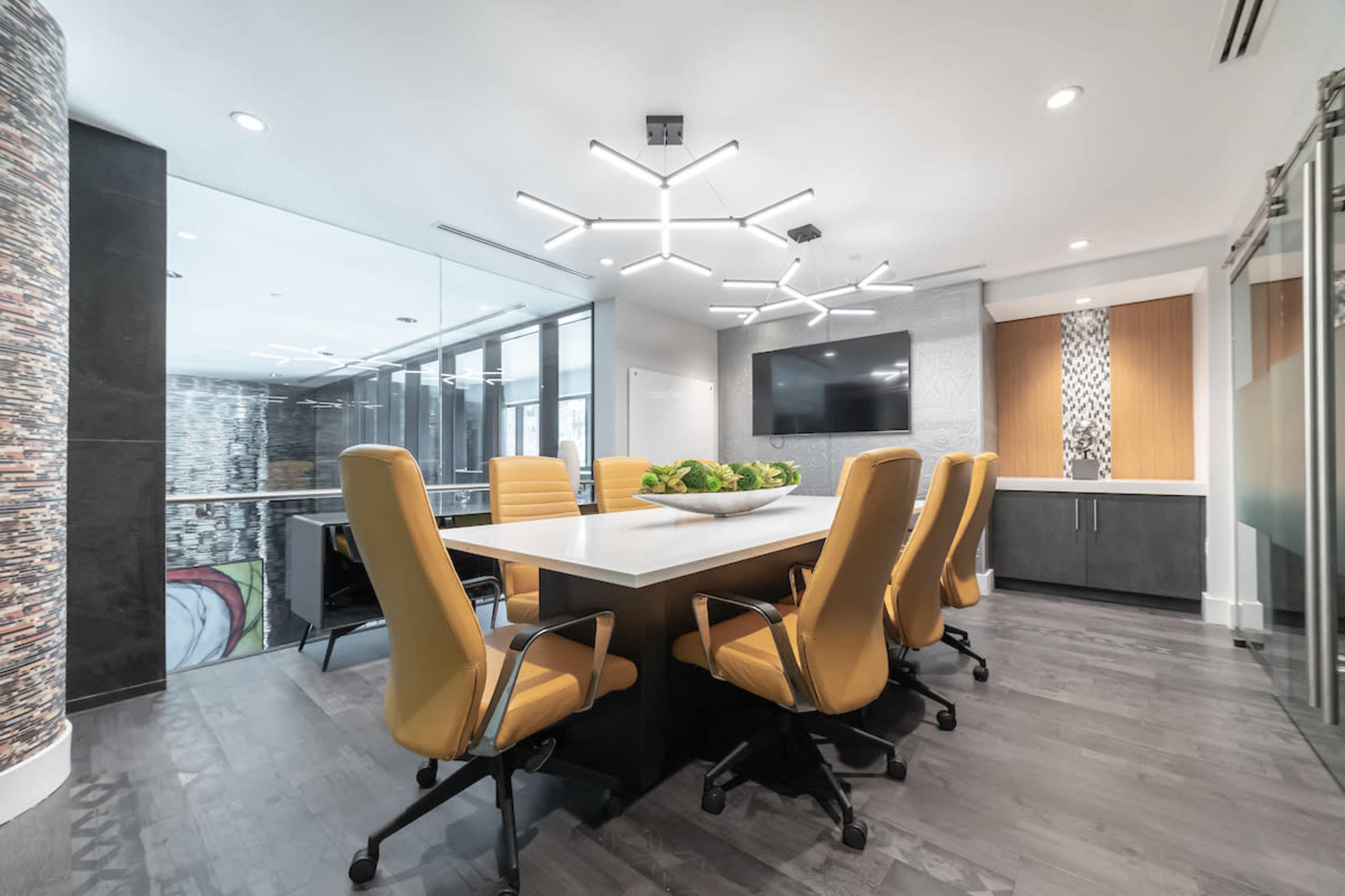 A modern conference room features a long table surrounded by yellow chairs and is illuminated by a contemporary light fixture, with a large window providing a view of the outside.