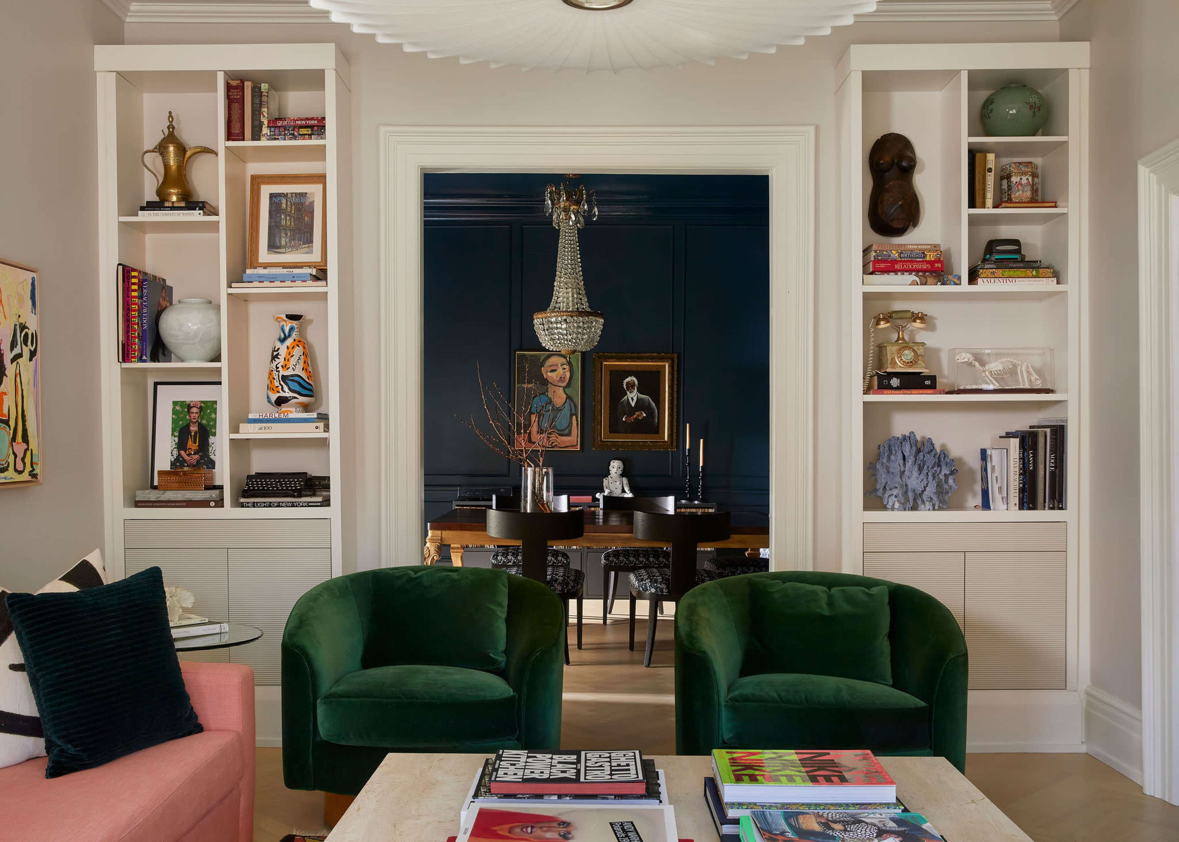 A stylish living room features two green armchairs, a pink sofa, and built-in shelves displaying decorative items and books, with a dark dining area visible through an archway.