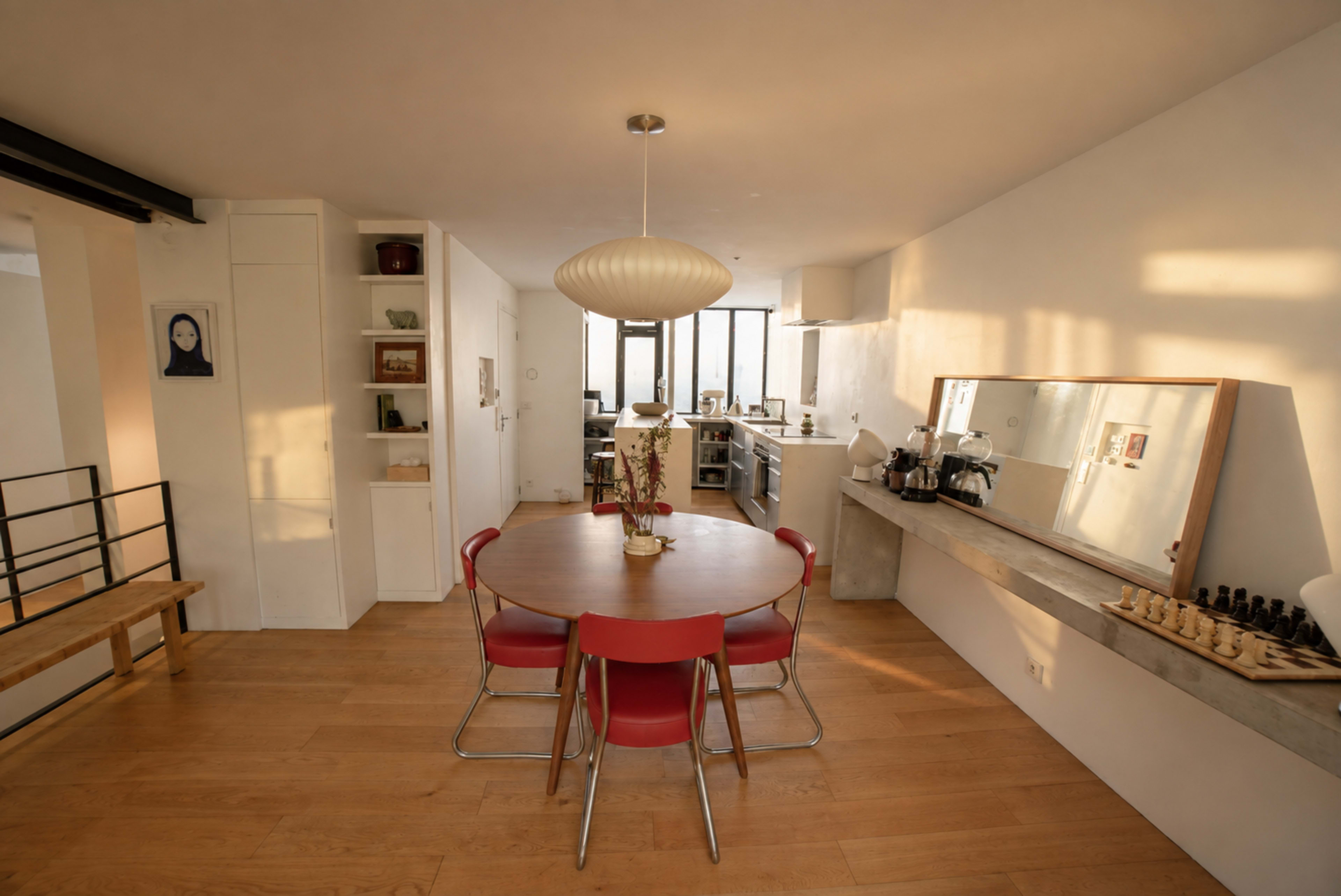 The image shows a modern dining area with a round table surrounded by red chairs, a minimalist kitchen in the background, and soft natural light illuminating the space.