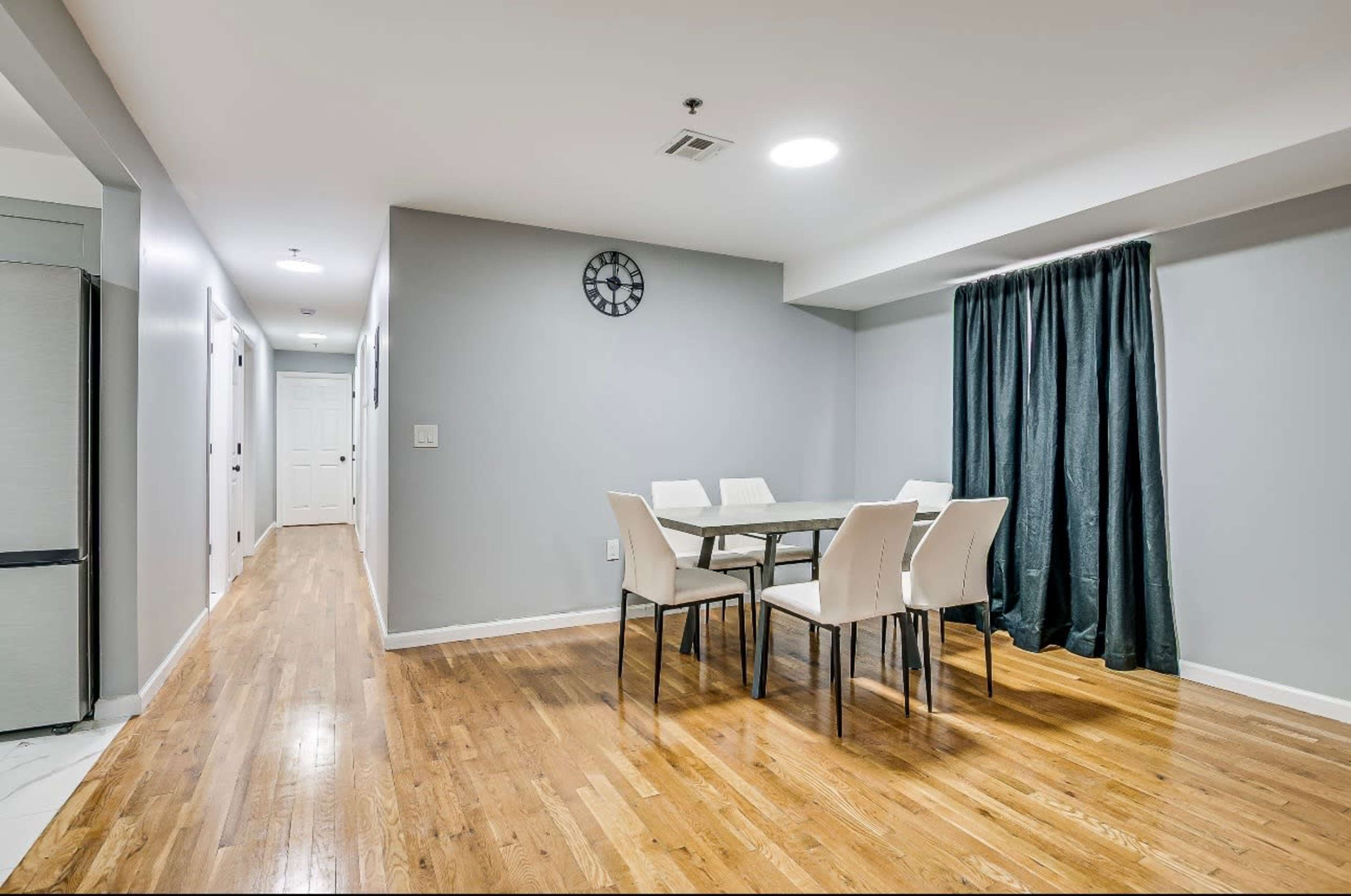 The image shows a dining area with a table and chairs, a wall clock, and a curtain, set within a corridor that leads to a door at the far end.