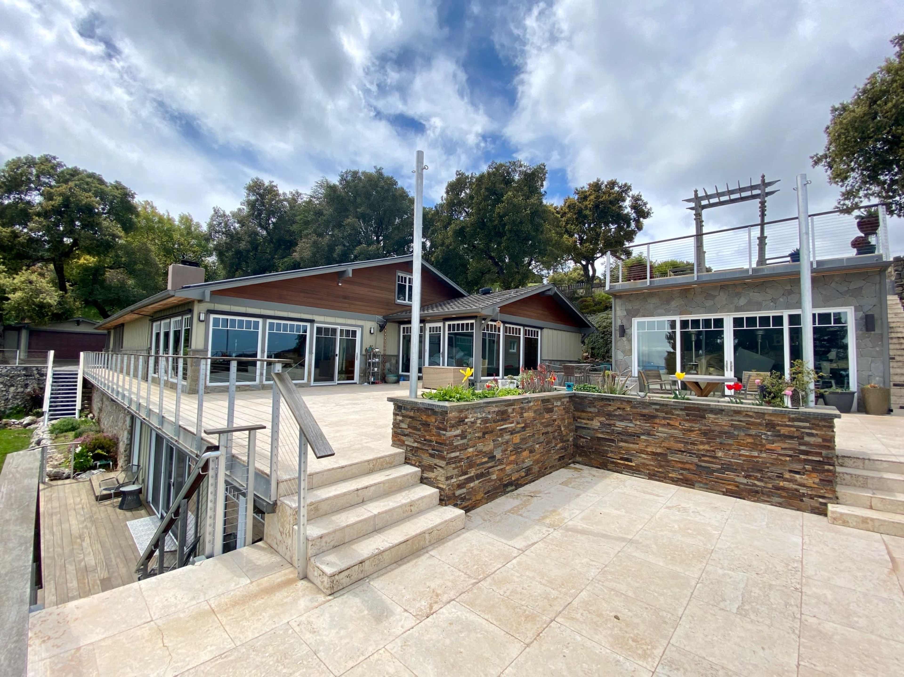 A modern two-story house with large windows, a stone patio, and steps leading down to a landscaped area surrounded by trees.