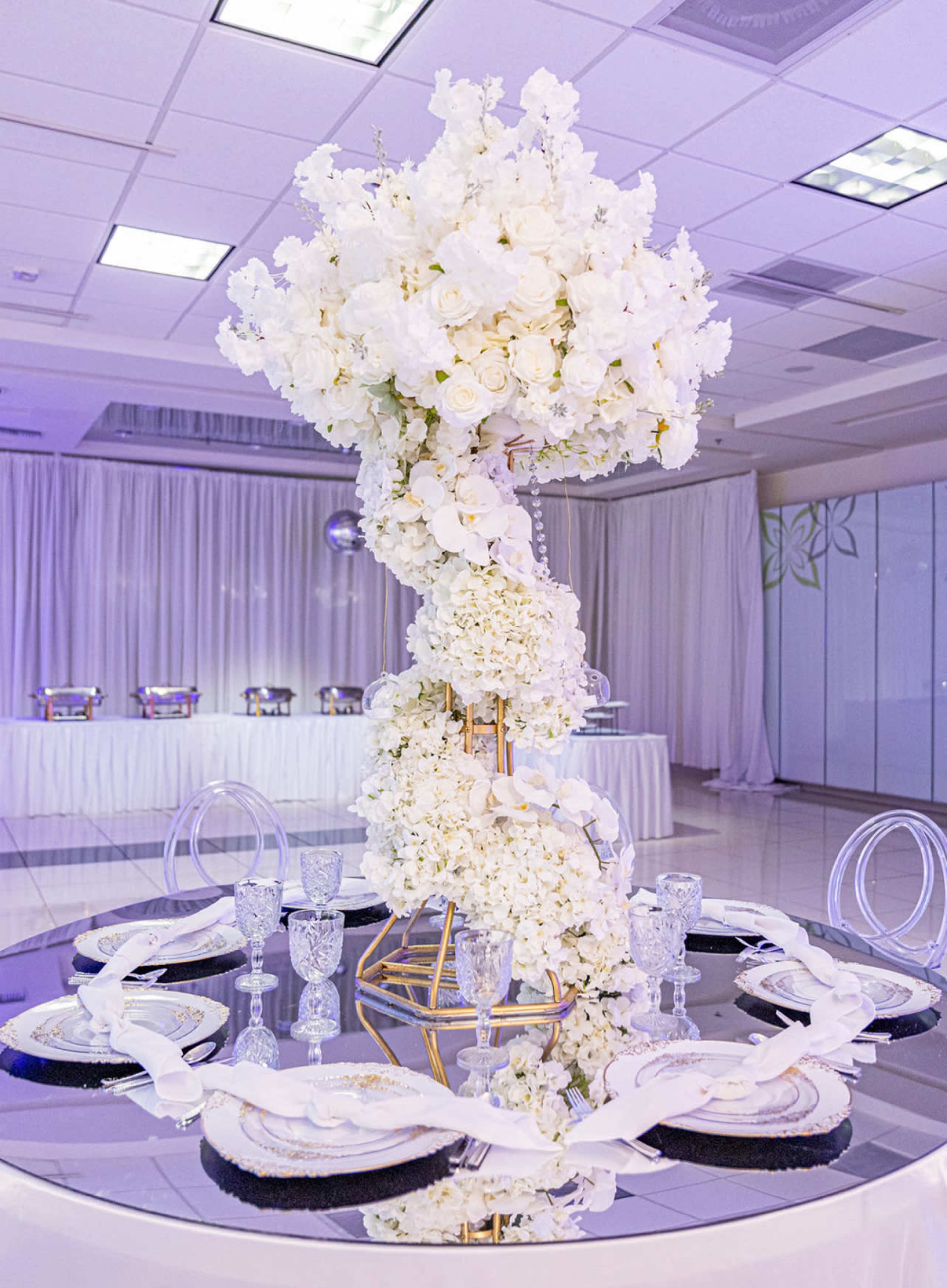 A tall floral centerpiece made of white flowers is displayed on a mirrored table surrounded by elegant place settings and crystal glassware in a banquet hall.