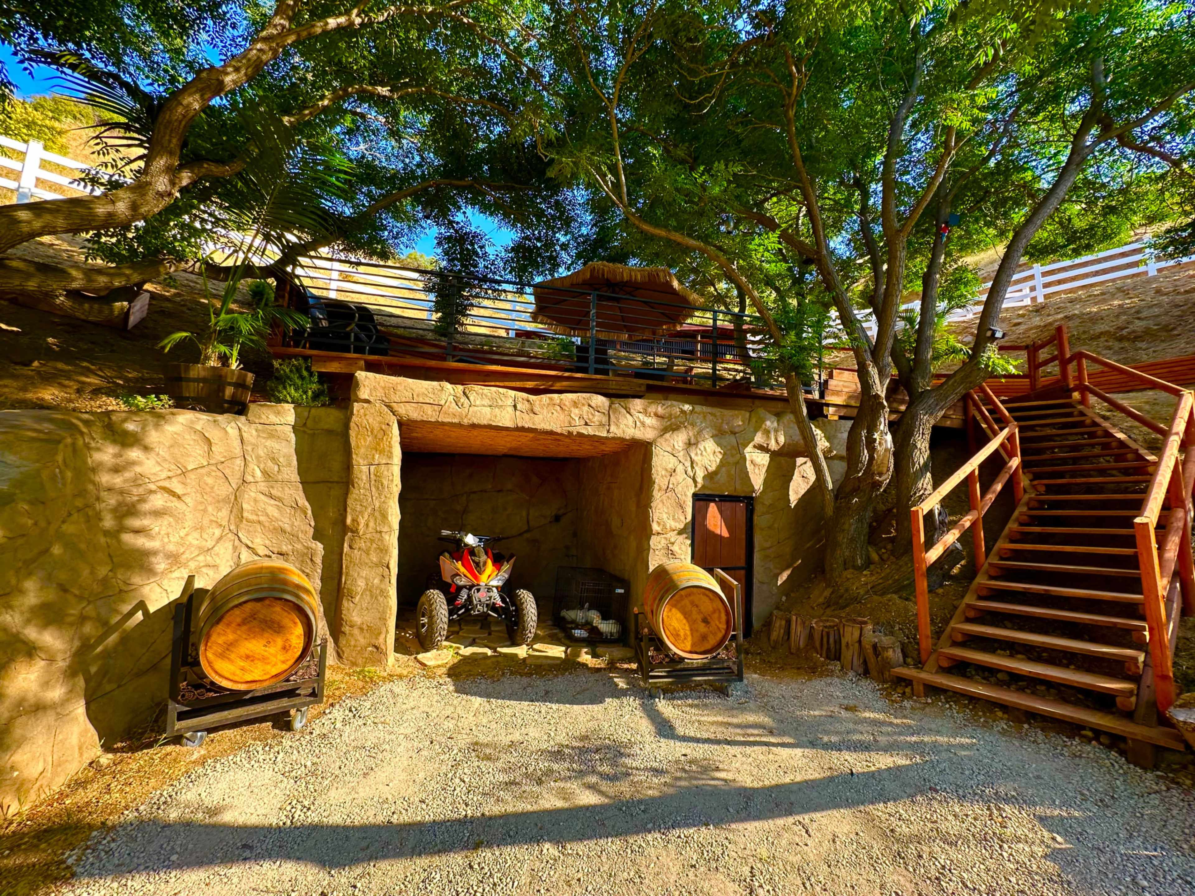 A rustic outdoor area featuring a stone entrance, wooden barrels, an ATV parked nearby, and a staircase leading up through a tree-lined landscape.