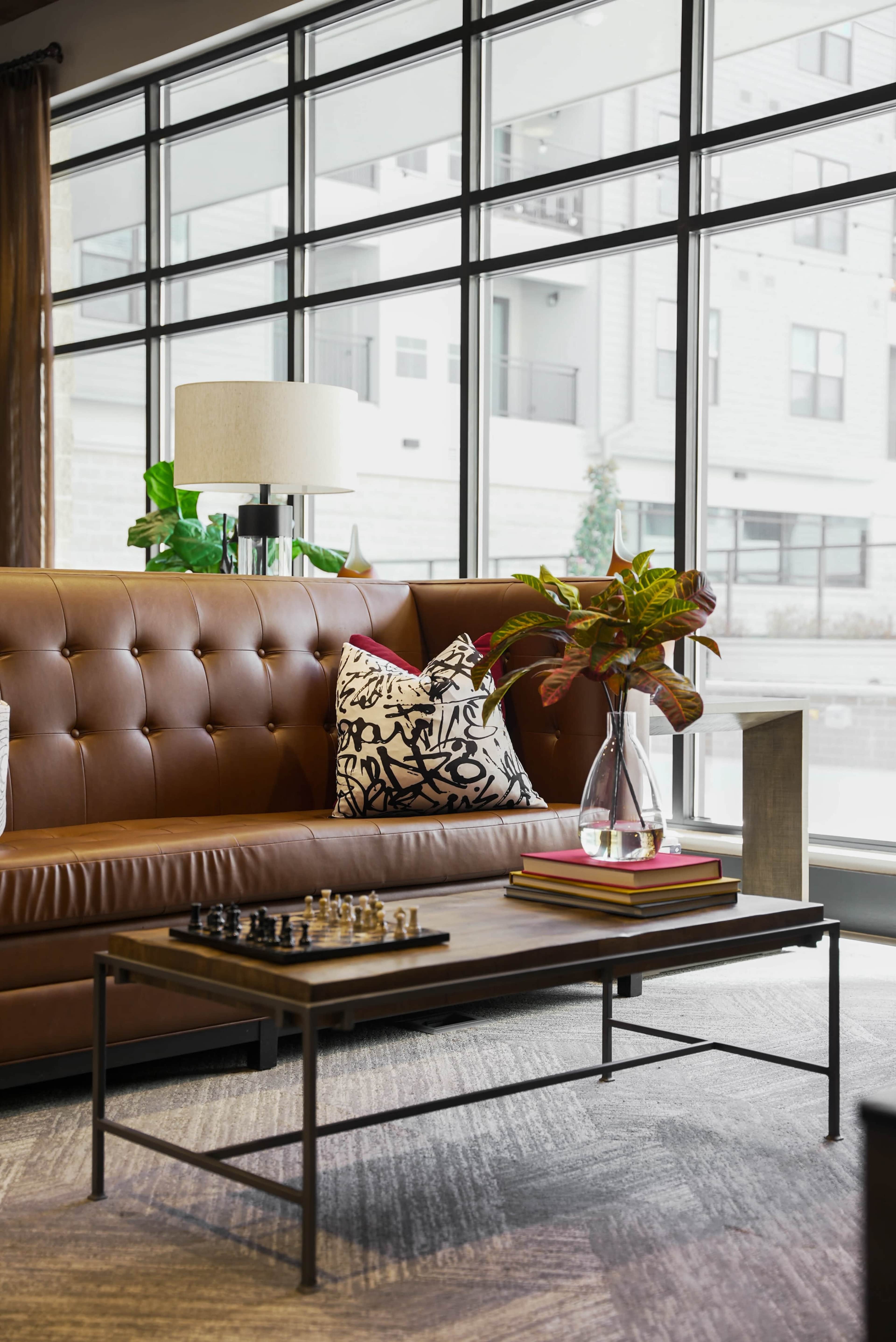 A modern living room features a brown tufted sofa, a decorative pillow, a glass coffee table with a chess set, and large windows providing ample natural light.