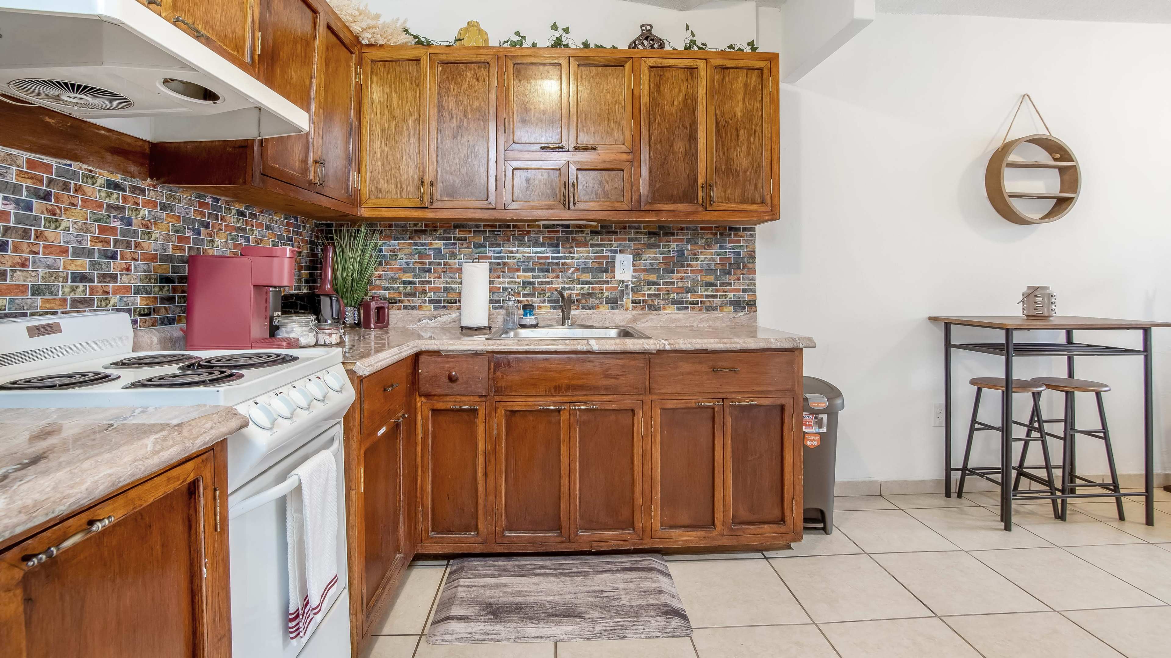 A compact kitchen with wooden cabinets, a tiled backsplash, and a small dining table with stools.