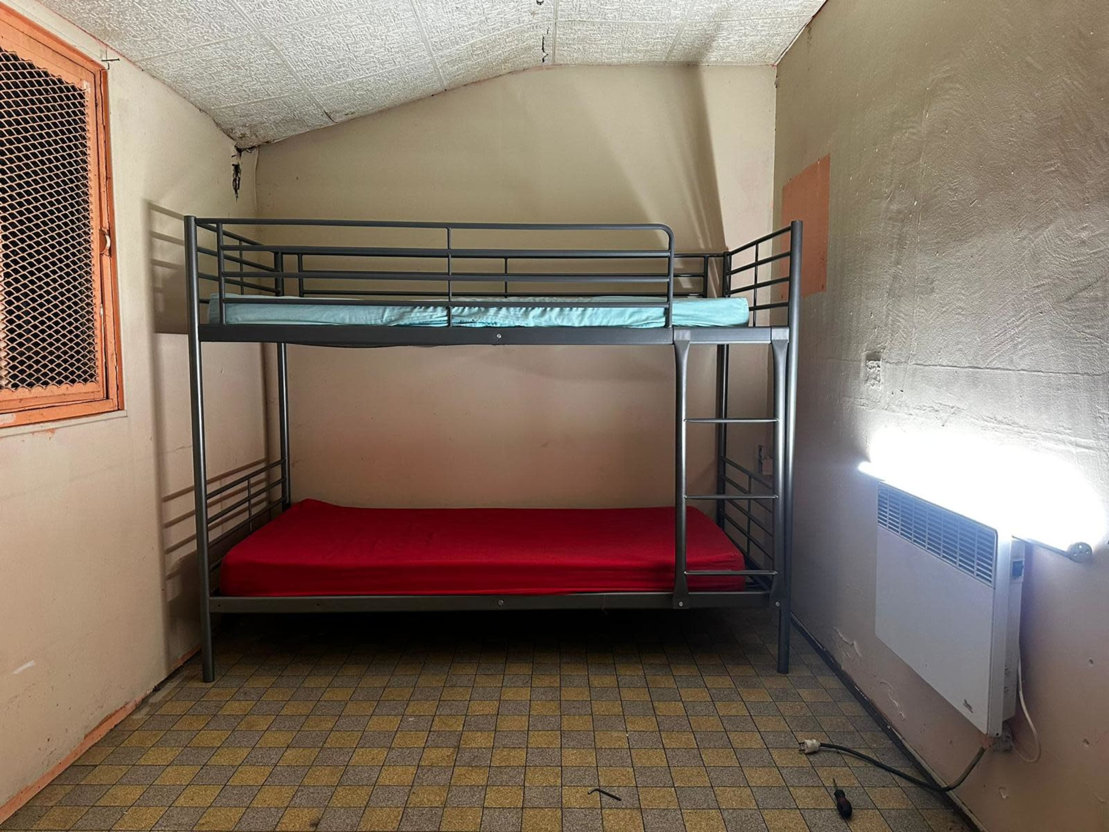 The image shows a small bedroom with a bunk bed, featuring a blue mattress on the top and a red mattress on the bottom, against a plain, light-colored wall.
