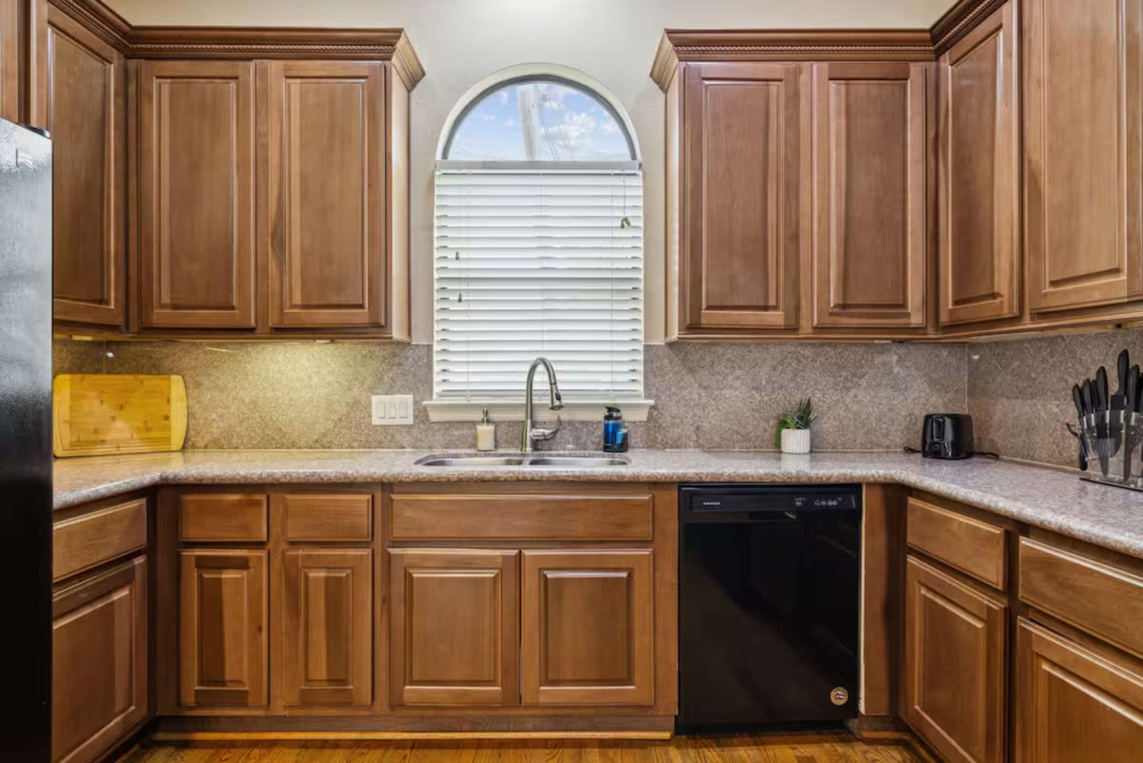 The image shows a modern kitchen featuring wooden cabinets, a granite countertop, a window with blinds, and a black dishwasher next to a sink.