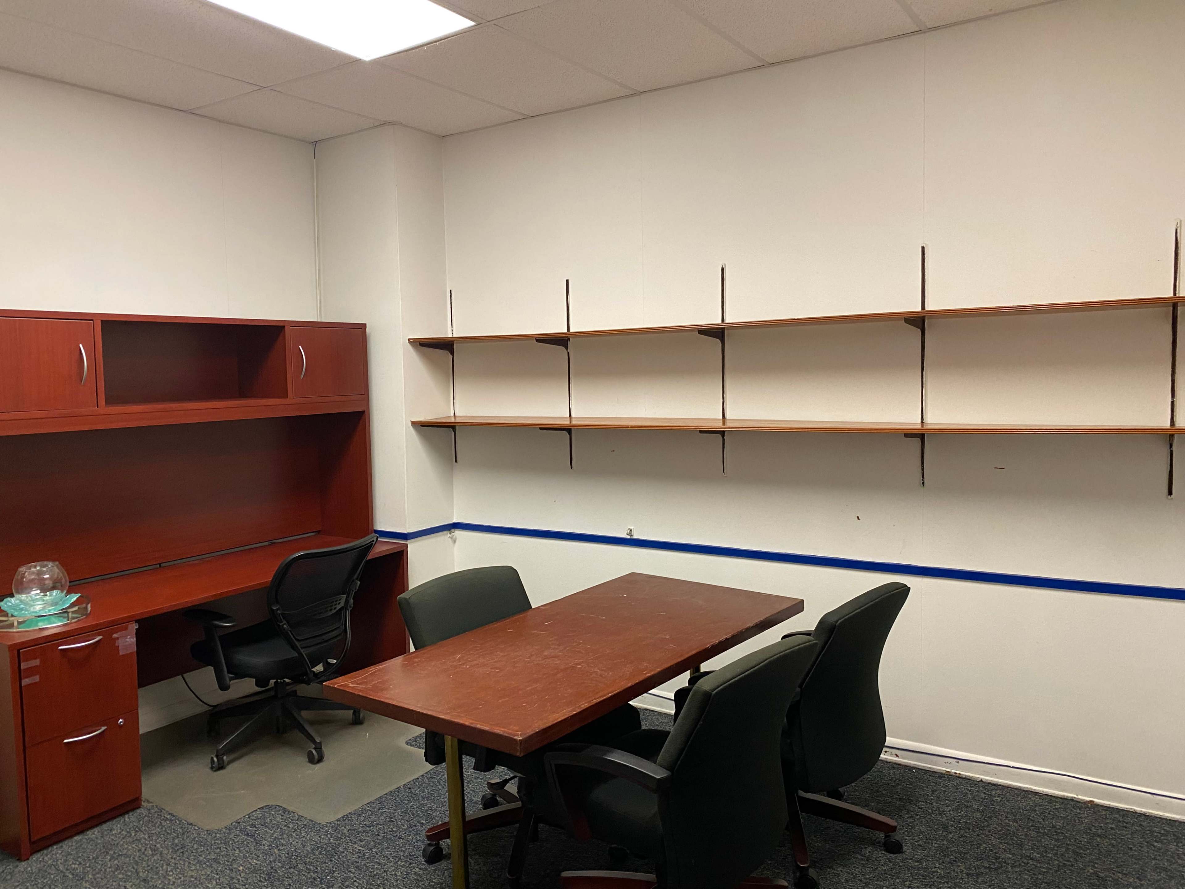 The image shows a small office space with a wooden desk, four chairs, and empty shelves on the wall.