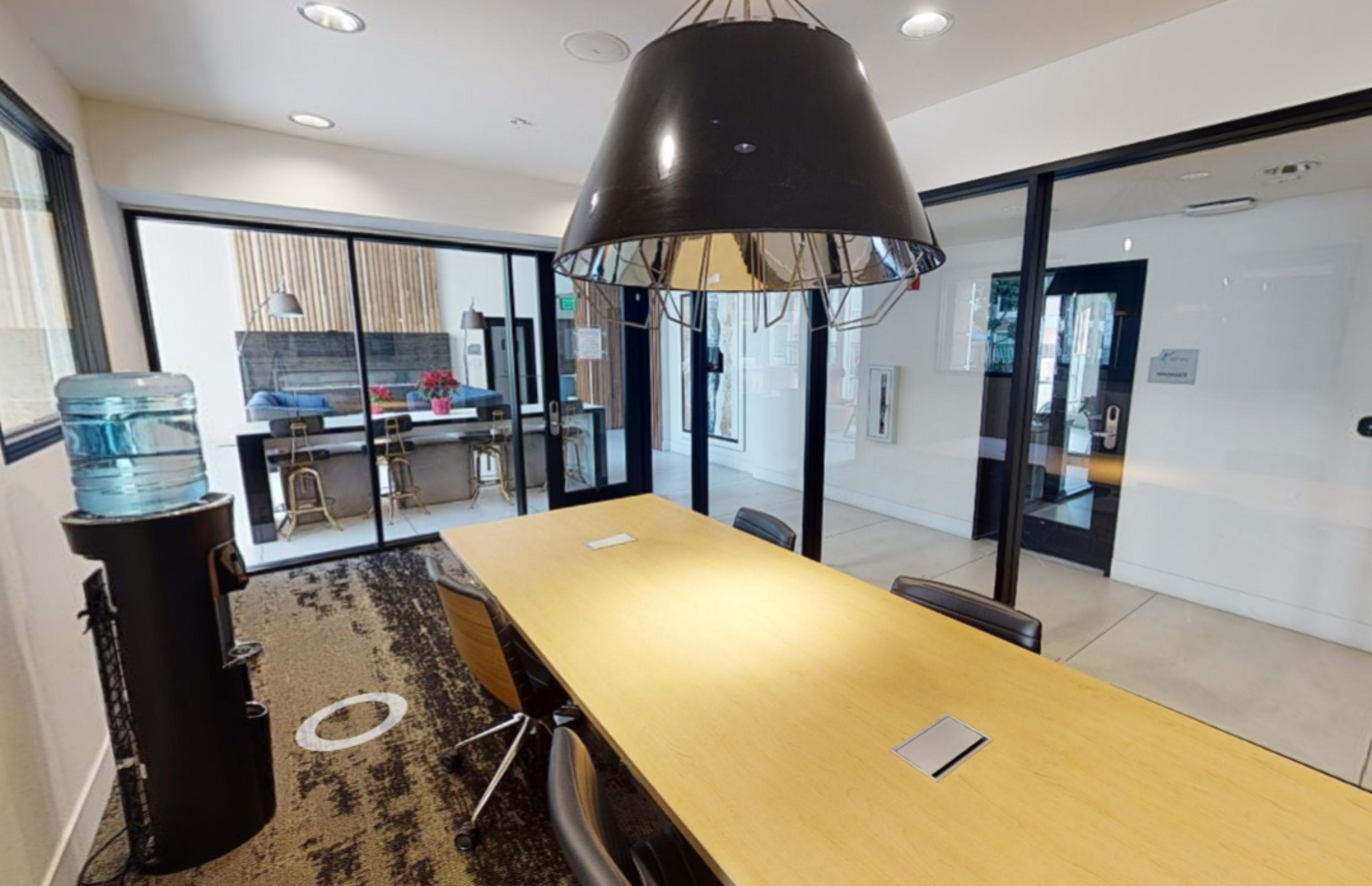 A modern conference room with a large wooden table, black chairs, and a water cooler, featuring glass walls that overlook an adjacent workspace.