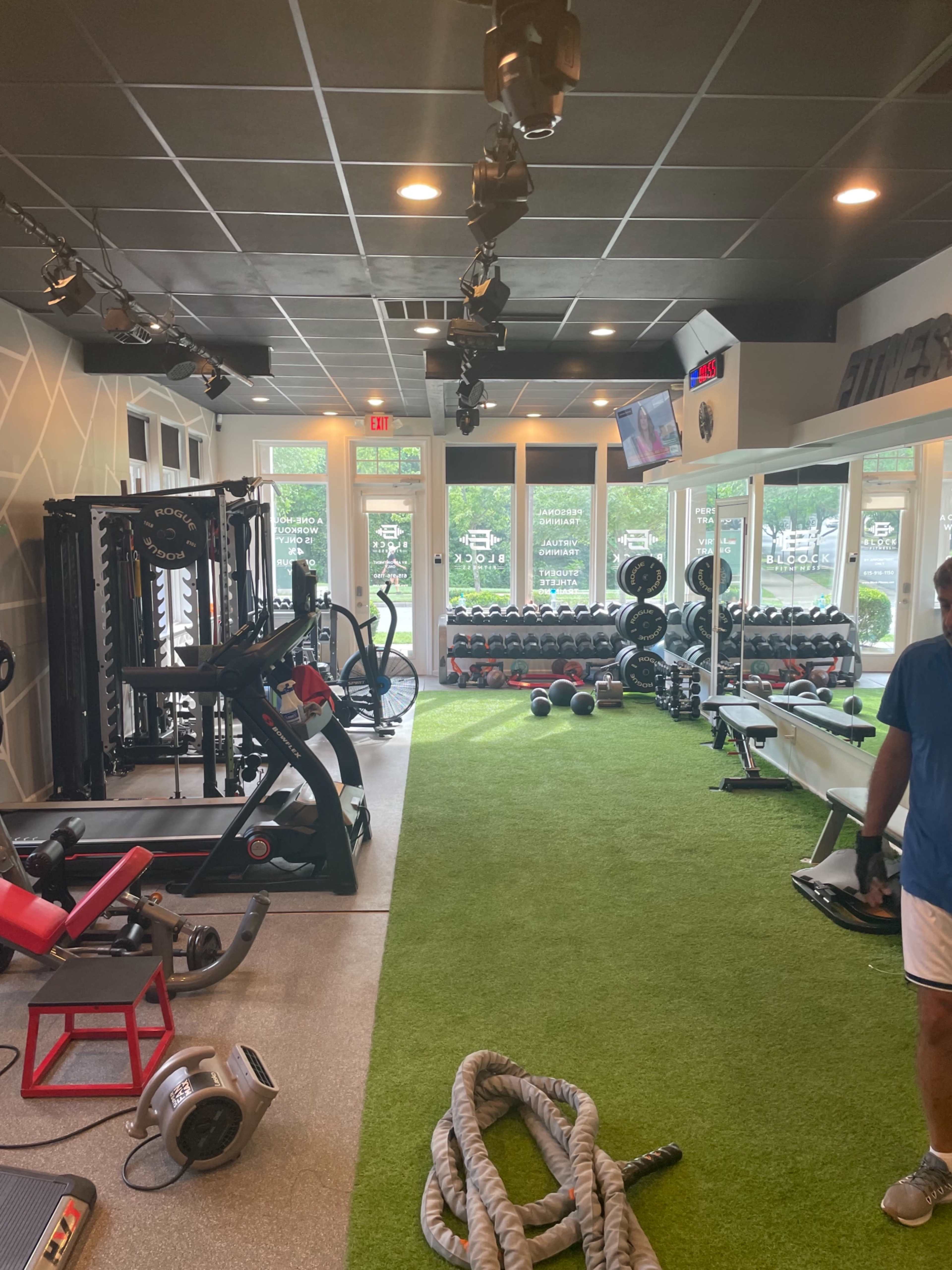 A modern gym interior featuring exercise equipment, weight racks, and green turf flooring.