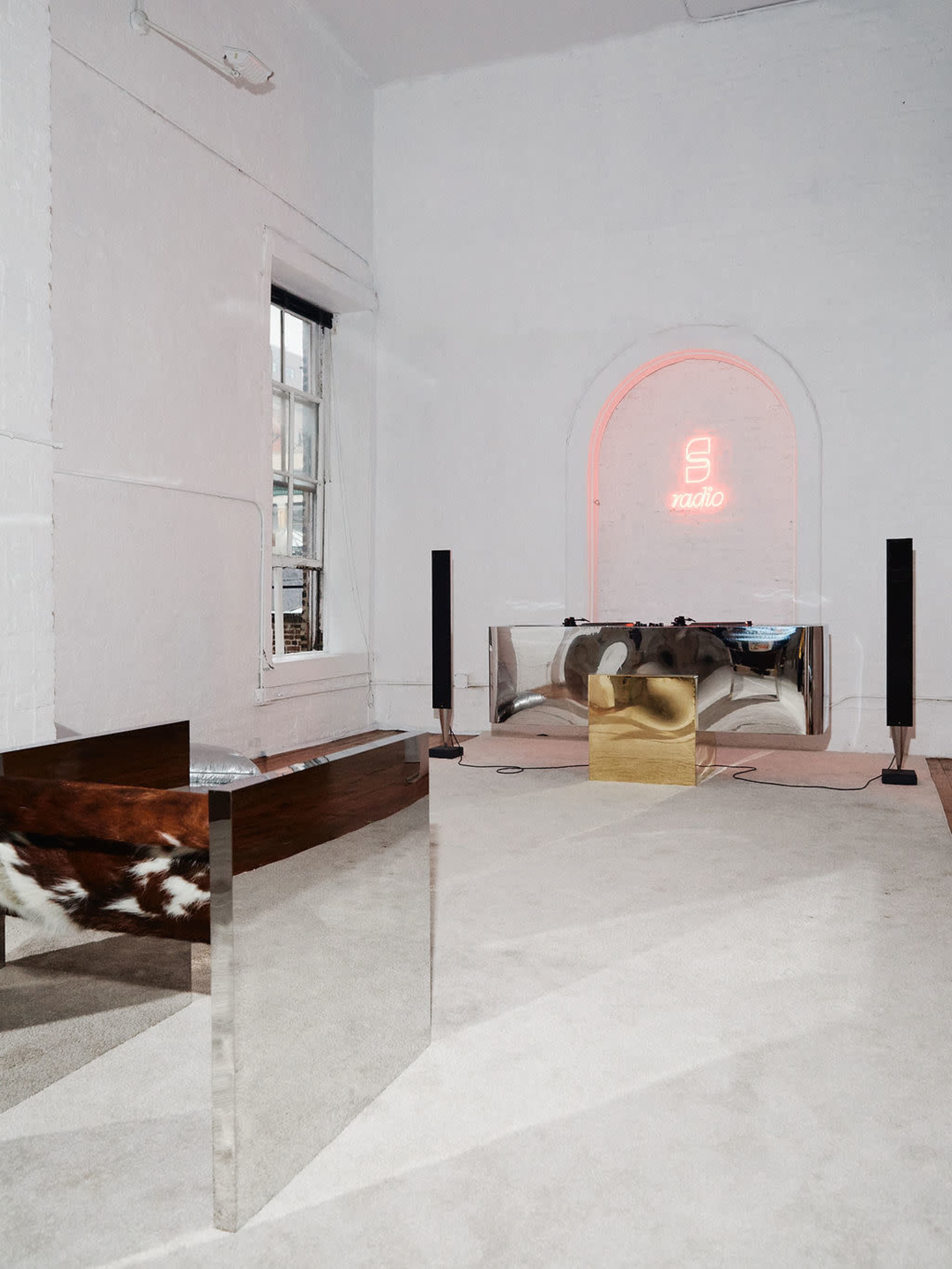 The image depicts a minimalist room featuring a silver and wooden table, a golden object, and a neon sign on the wall.