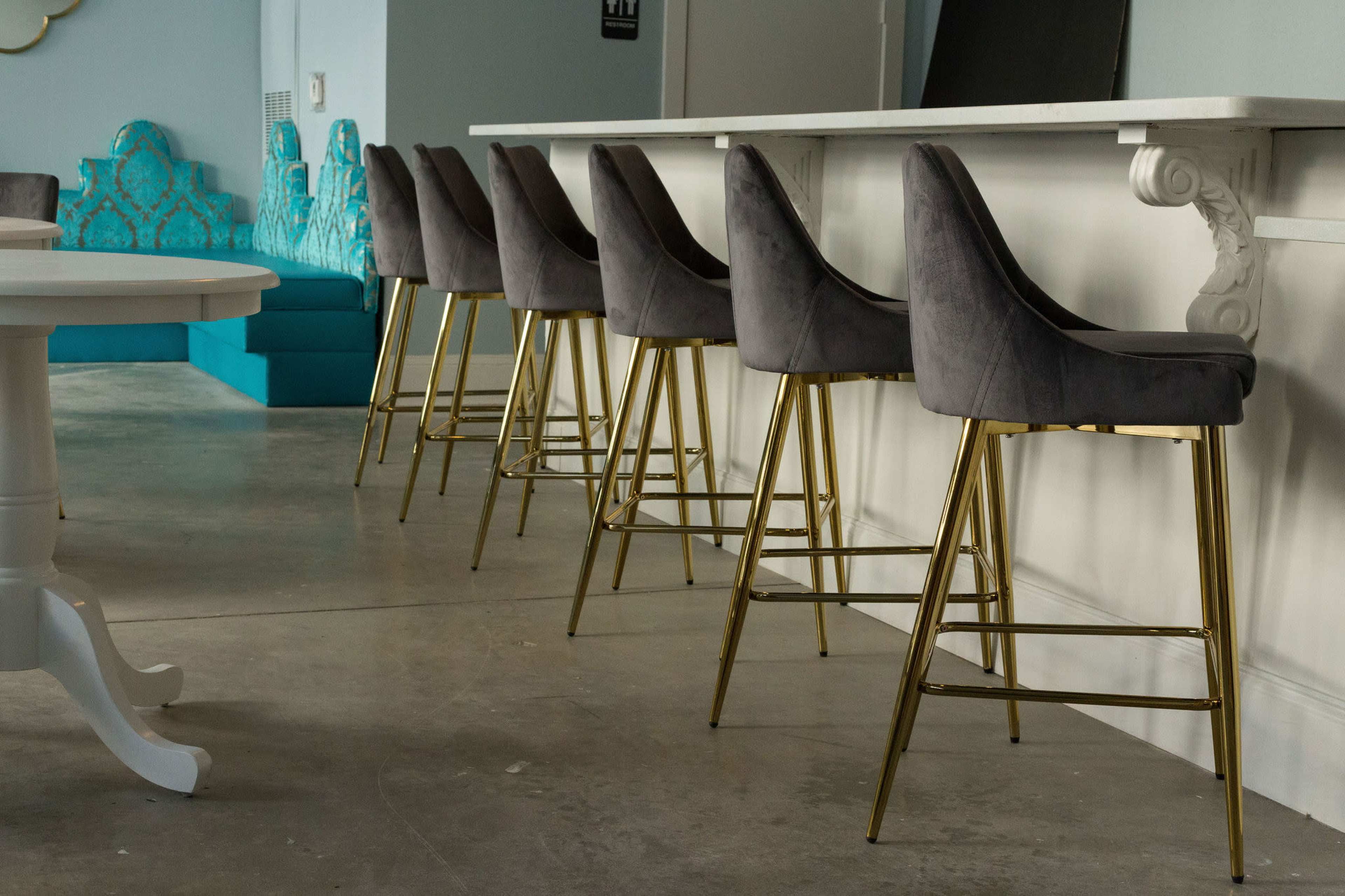 A row of gray upholstered bar stools with gold legs are positioned in front of a white countertop in a modern interior space.