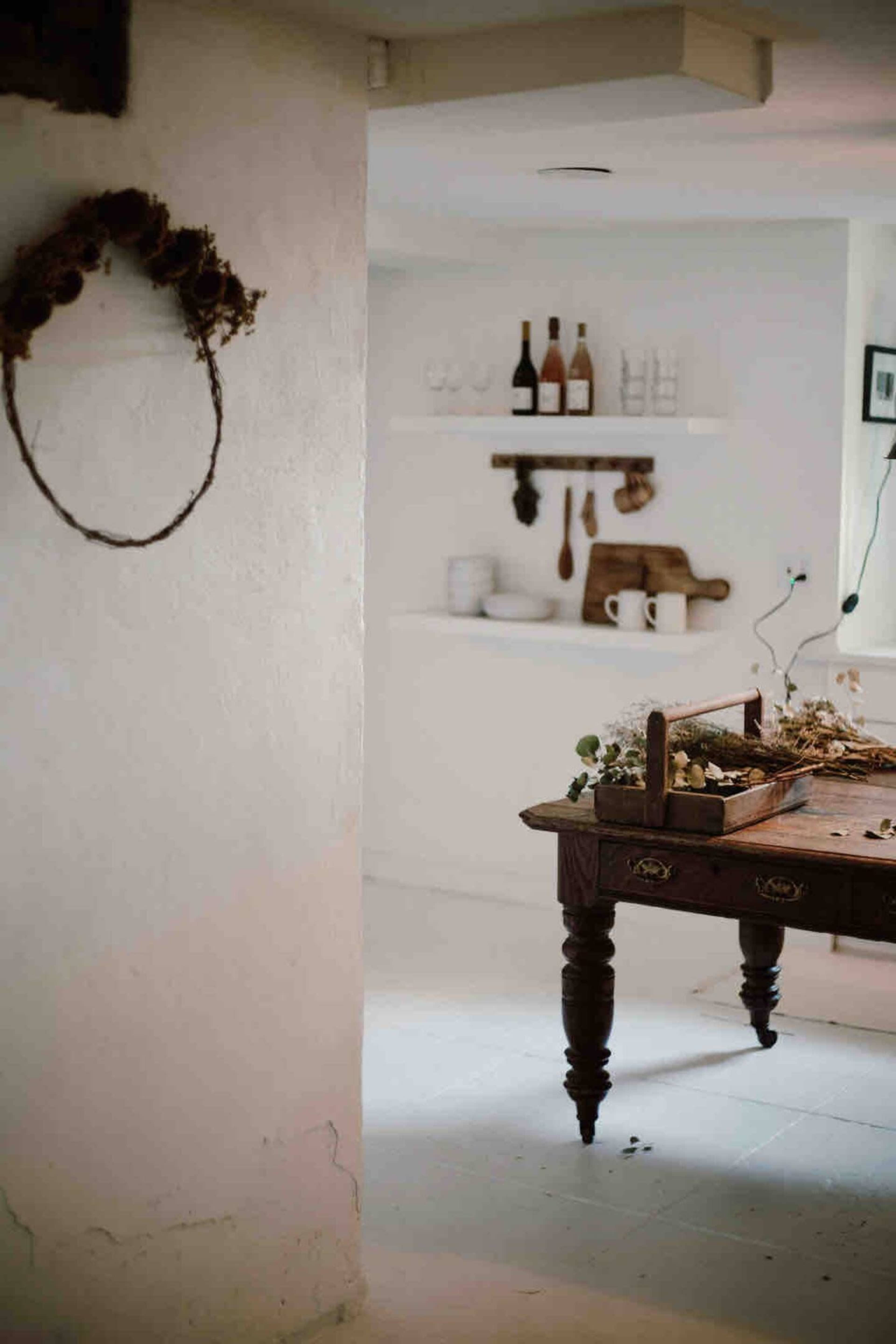 A wooden table with a rustic centerpiece is positioned in a softly lit kitchen that features white walls and open shelving with dishes and bottles.