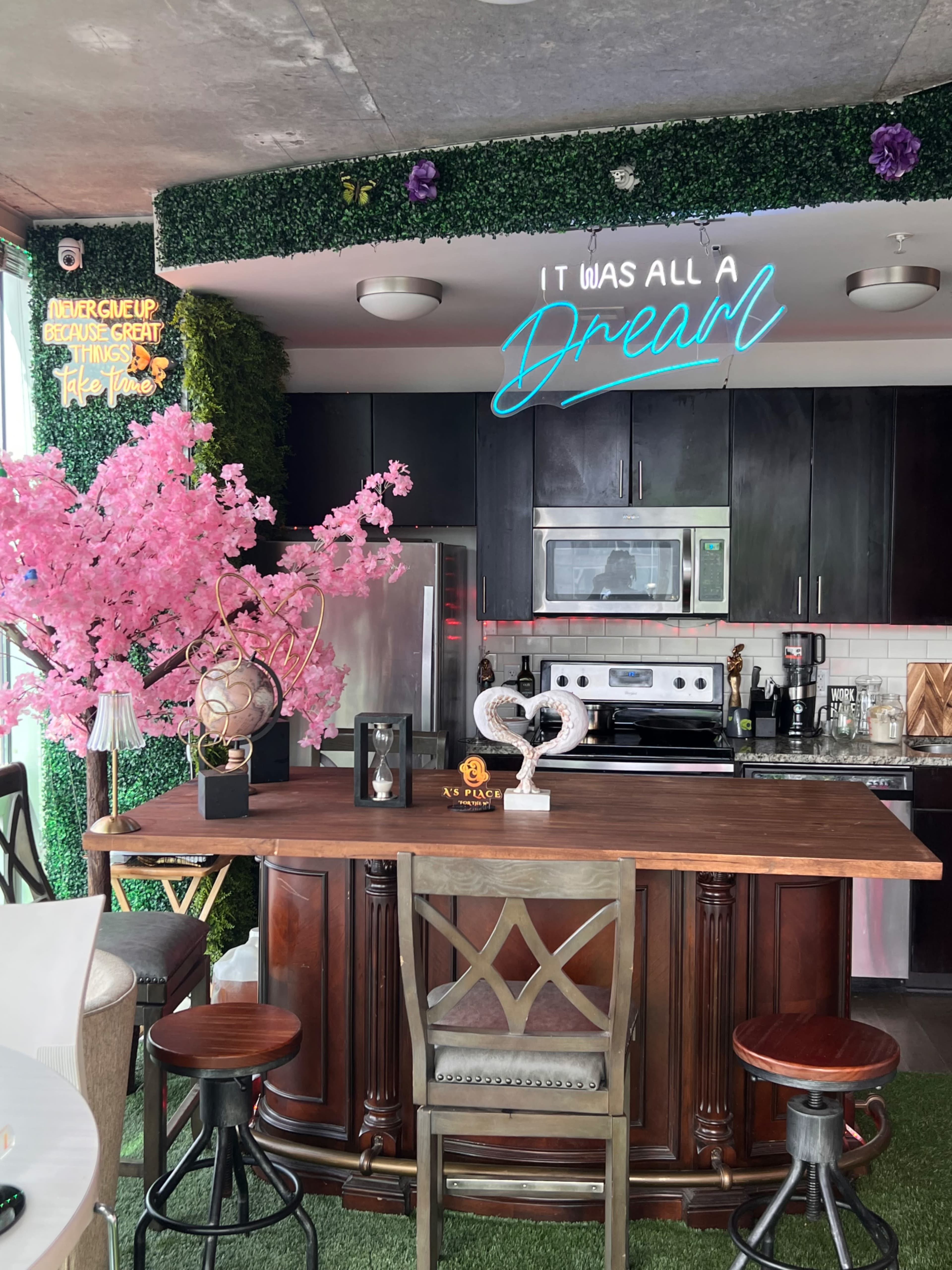 The image shows a modern kitchen with a wooden island, black cabinetry, a stainless steel refrigerator, and decorative elements like a pink cherry blossom tree and a neon sign that says, "It was all a Dream."