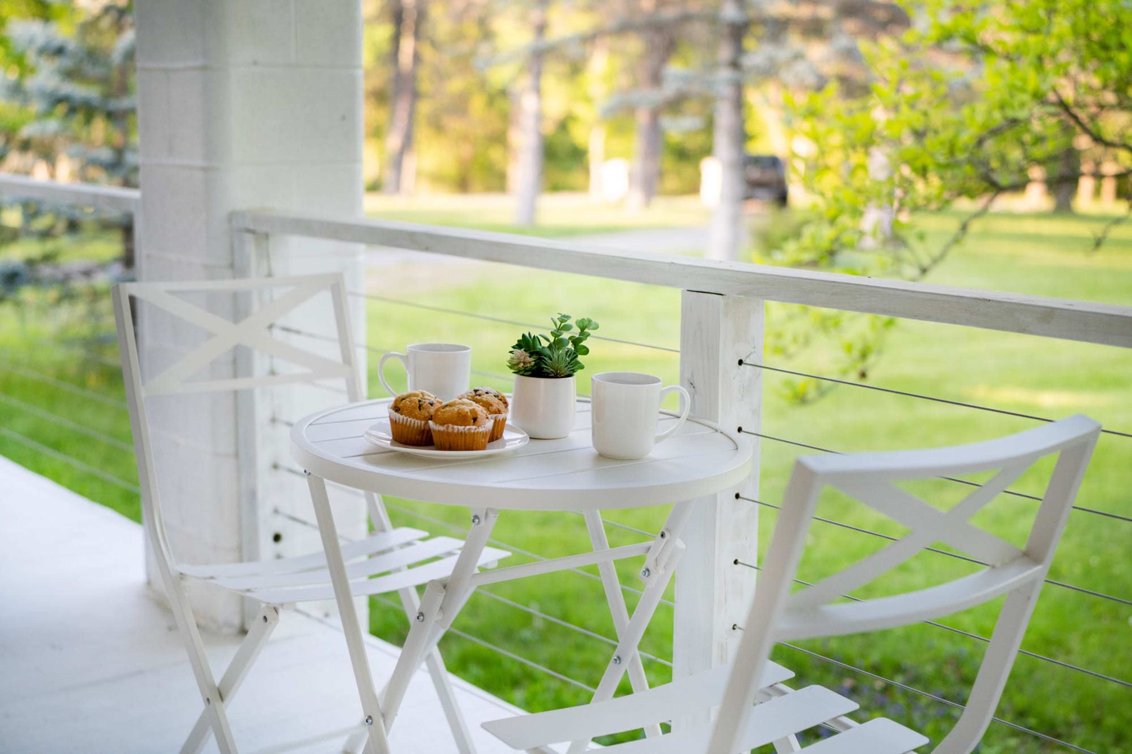 A small white table with two chairs holds a plate of muffins and two cups near a greenery-filled outdoor area.