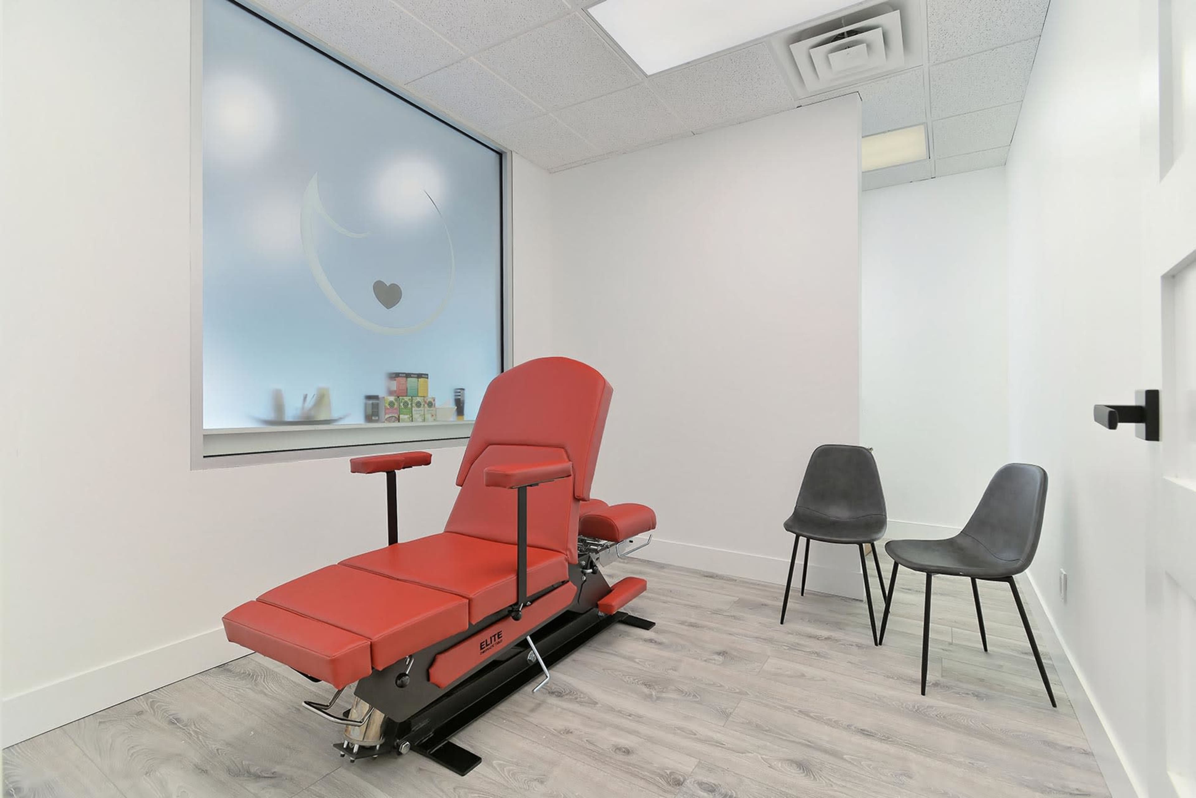A minimalist treatment room equipped with a red examination chair and two black chairs against a light-colored wall.
