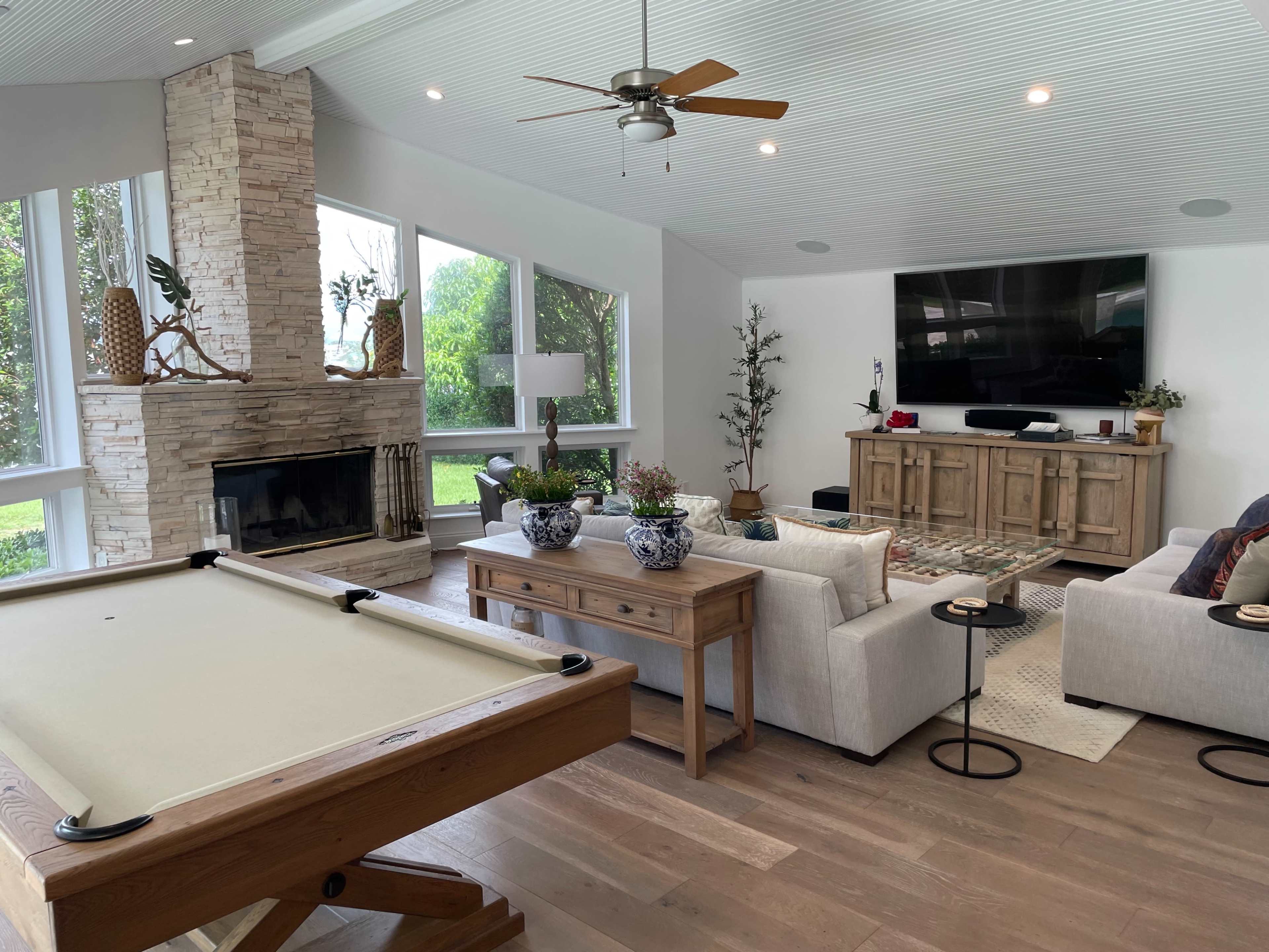 A modern living room with a pool table, a stone fireplace, large windows, and light-colored furniture.