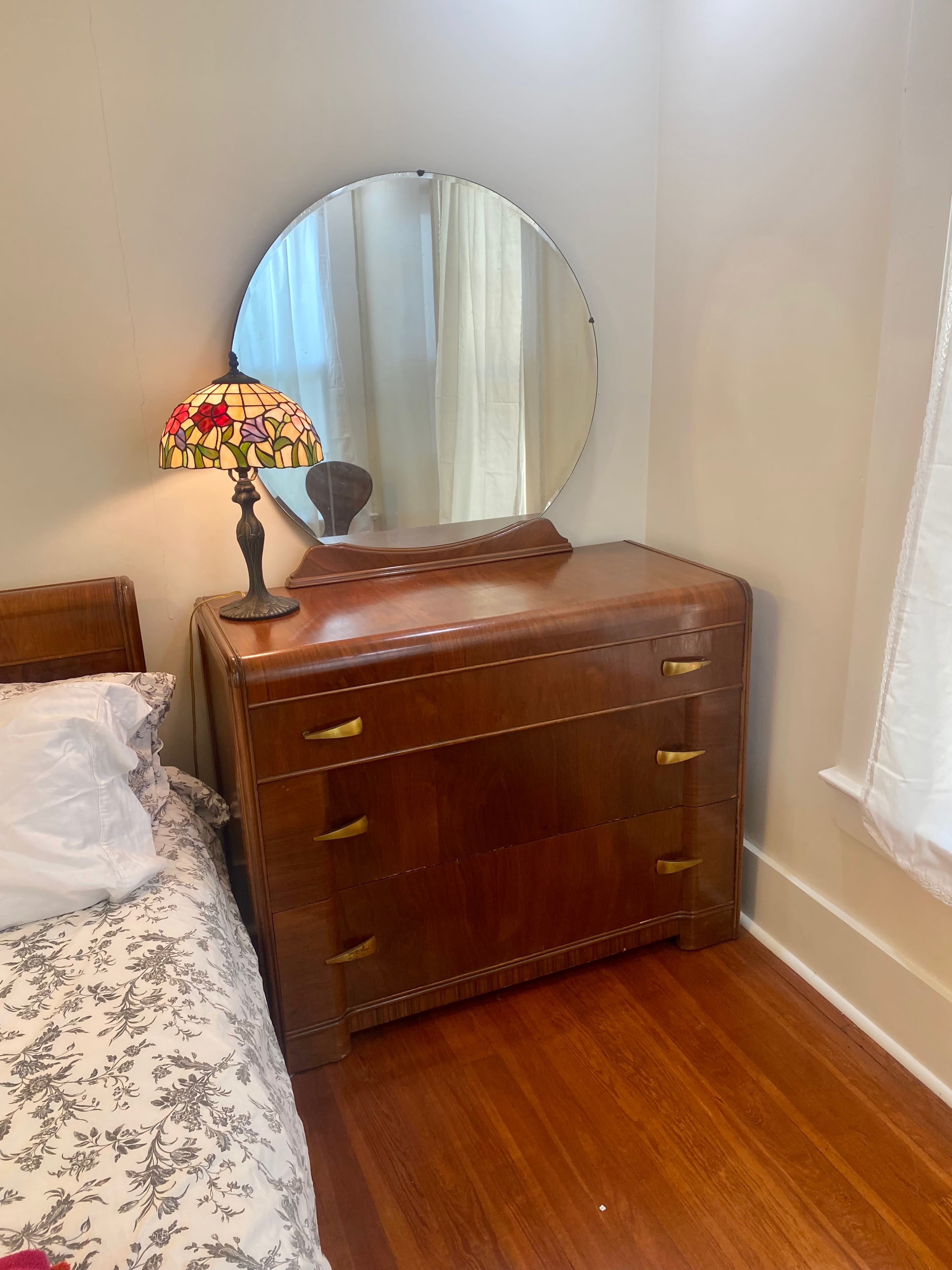 A wooden dresser with a round mirror above it, next to a bed with a floral-patterned blanket and a stained glass lamp beside the dresser.
