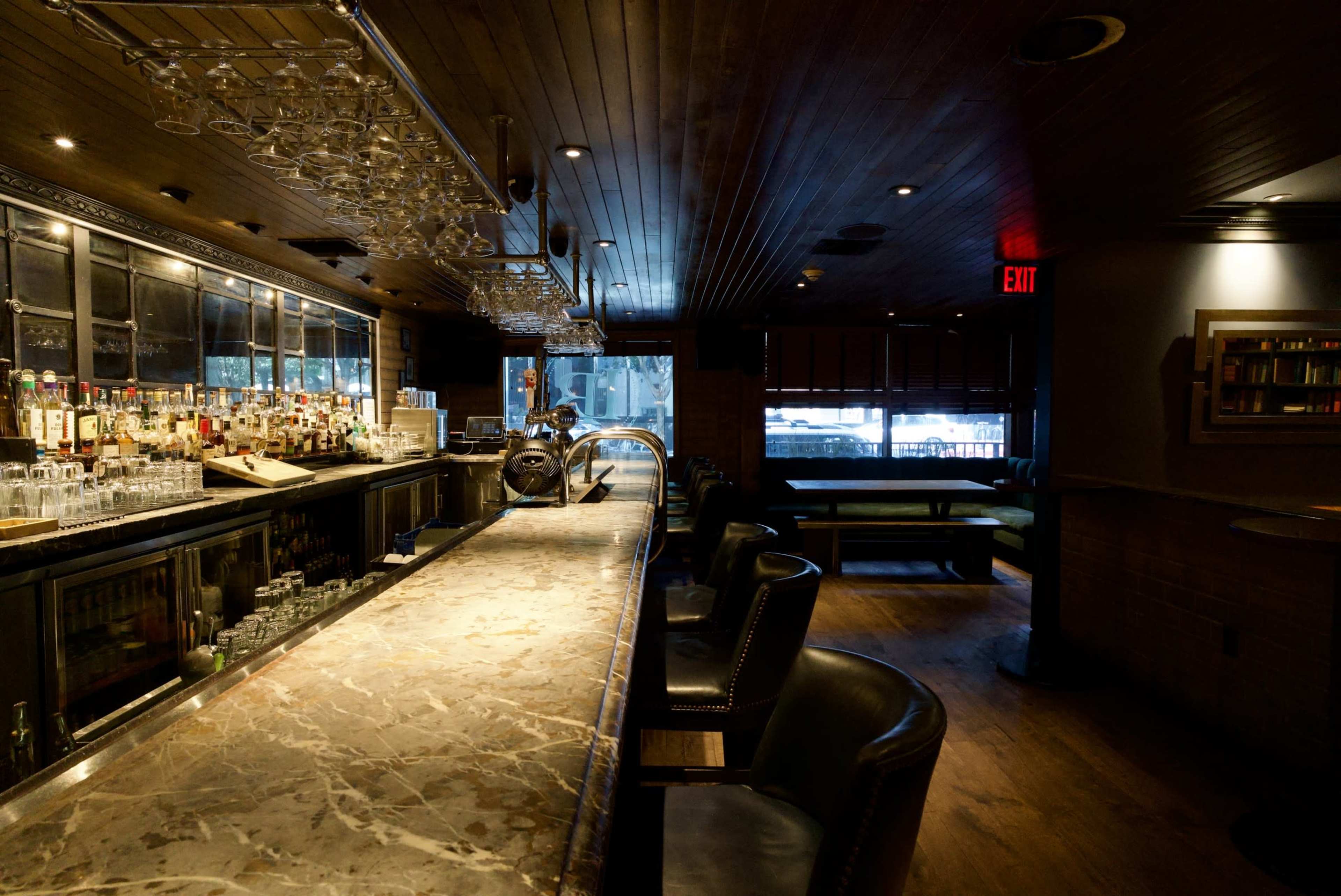 The image shows a dimly lit bar interior with a long marble countertop, shelves of liquor in the background, and a few booths along the sides.