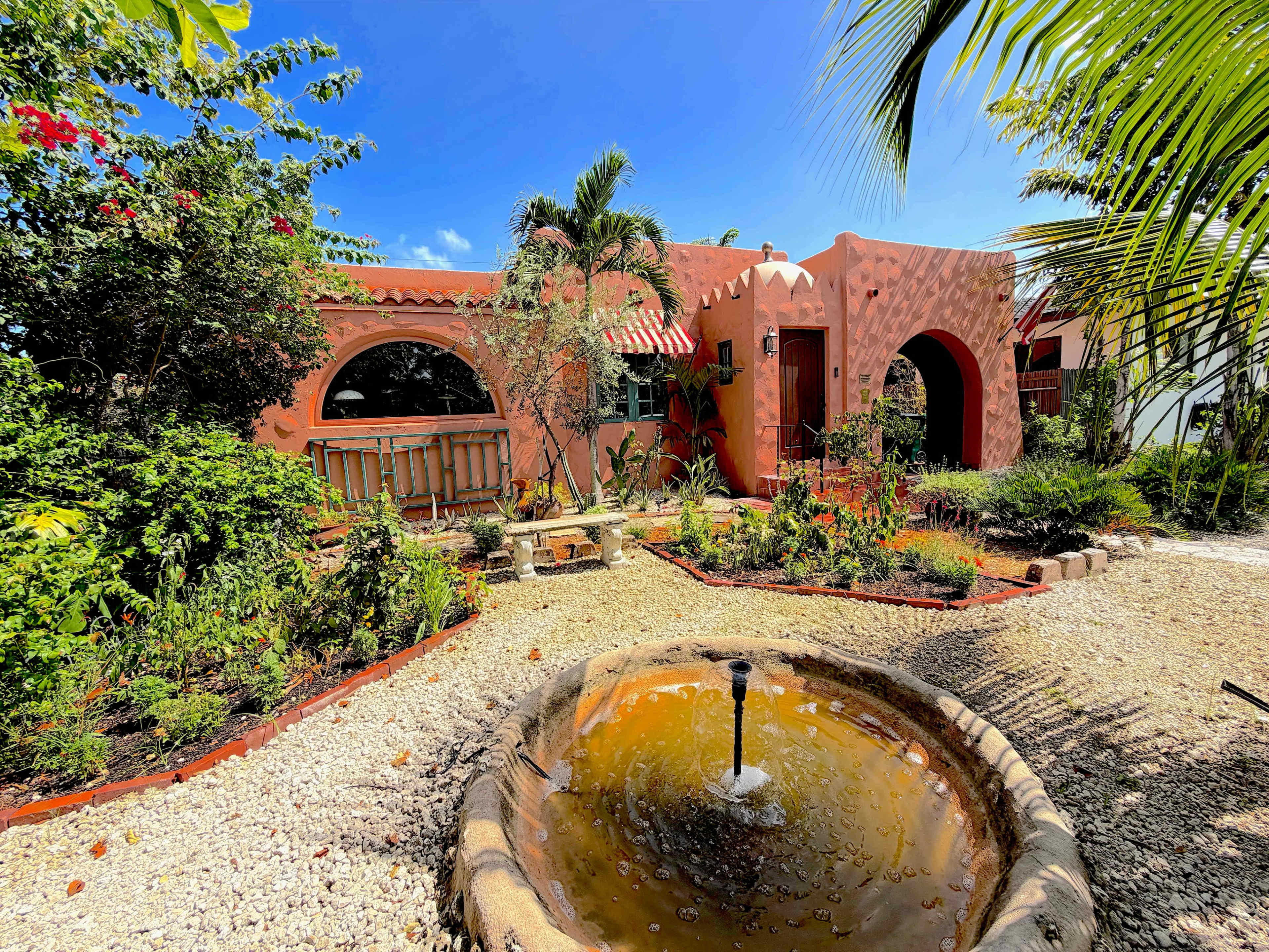 The image shows a colorful building surrounded by lush greenery and a circular fountain in a garden setting.