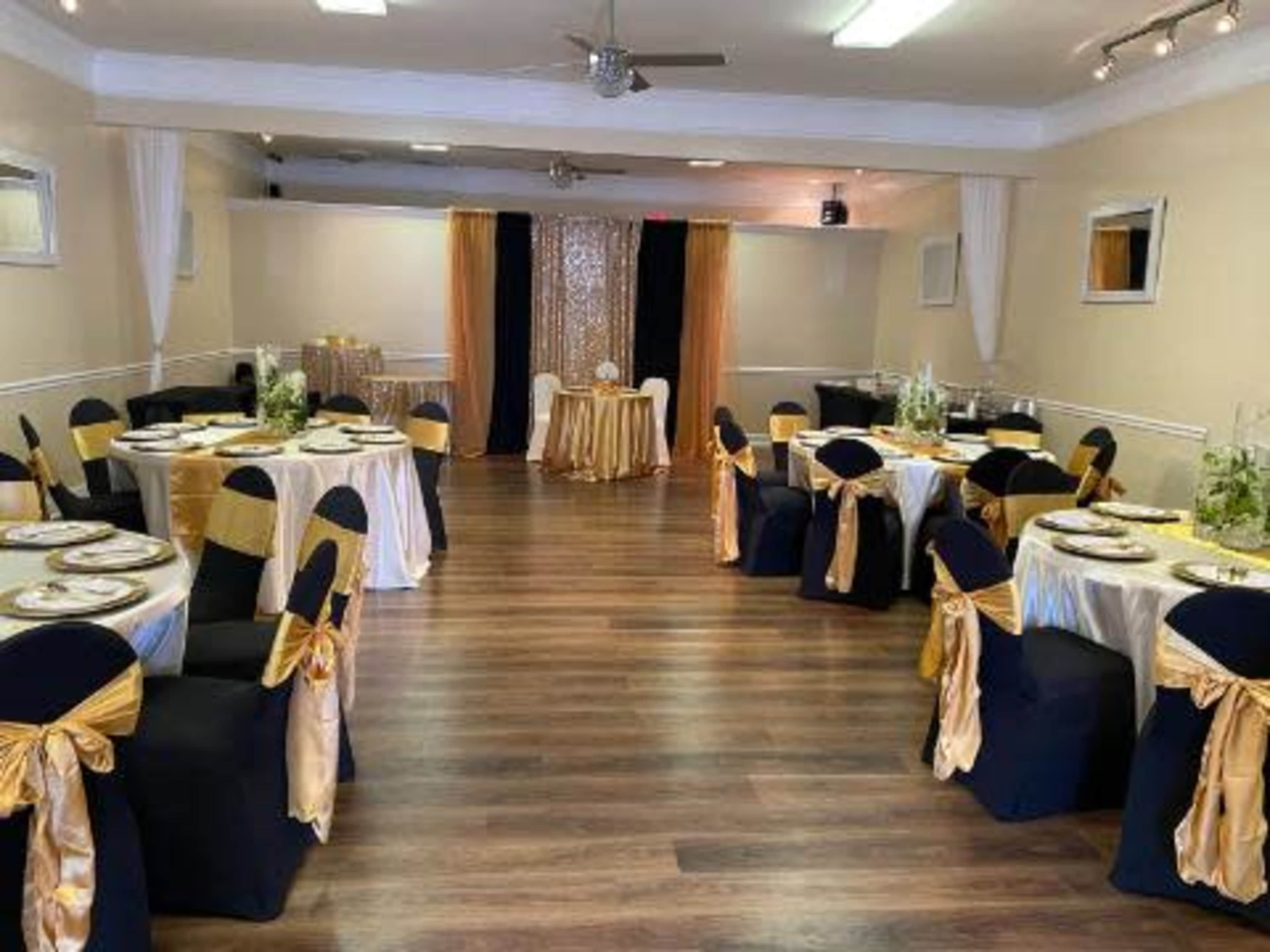 A banquet hall is set up with round tables covered in white and gold linens, surrounded by black chair covers with gold sashes.