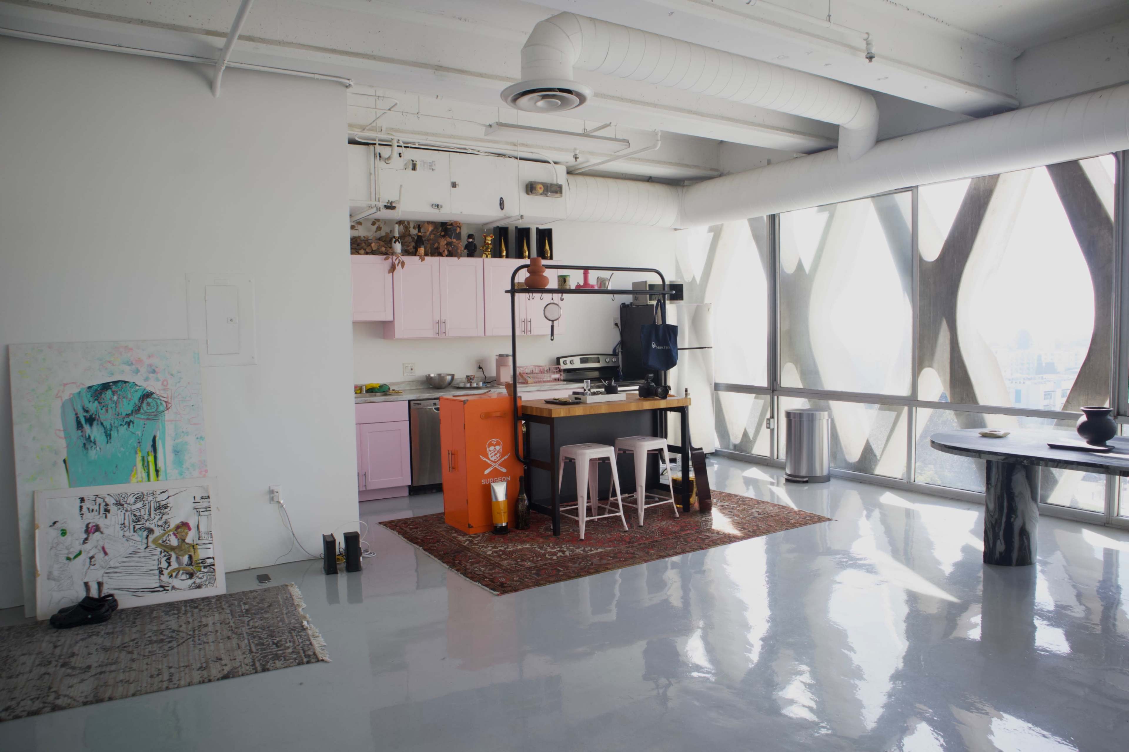 A modern kitchen and dining area featuring pink cabinetry, an orange refrigerator, and large windows with a city view.