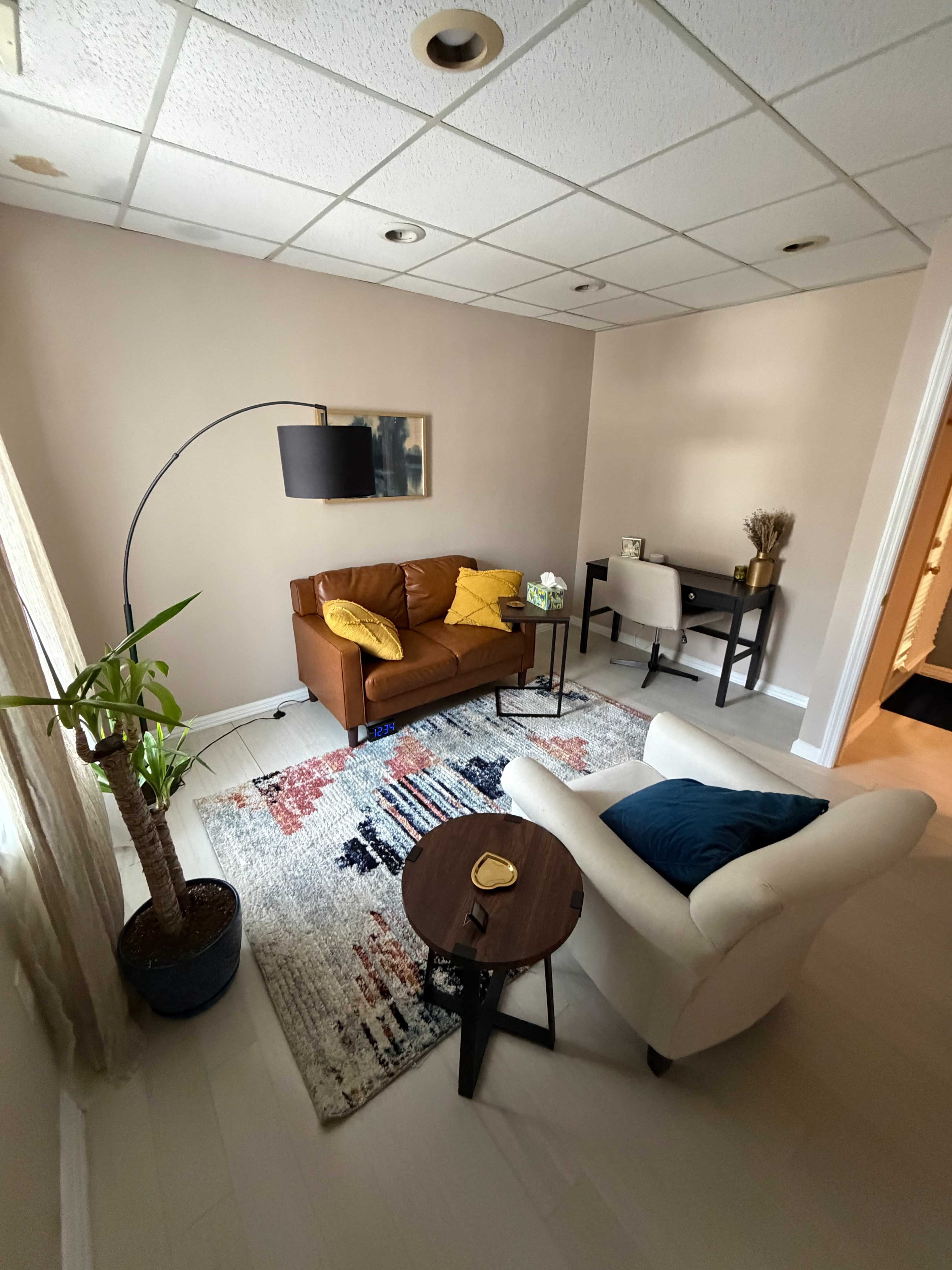 The image shows a cozy living room with a brown leather sofa, a small round coffee table, a desk, and a potted plant, all set on a patterned rug.
